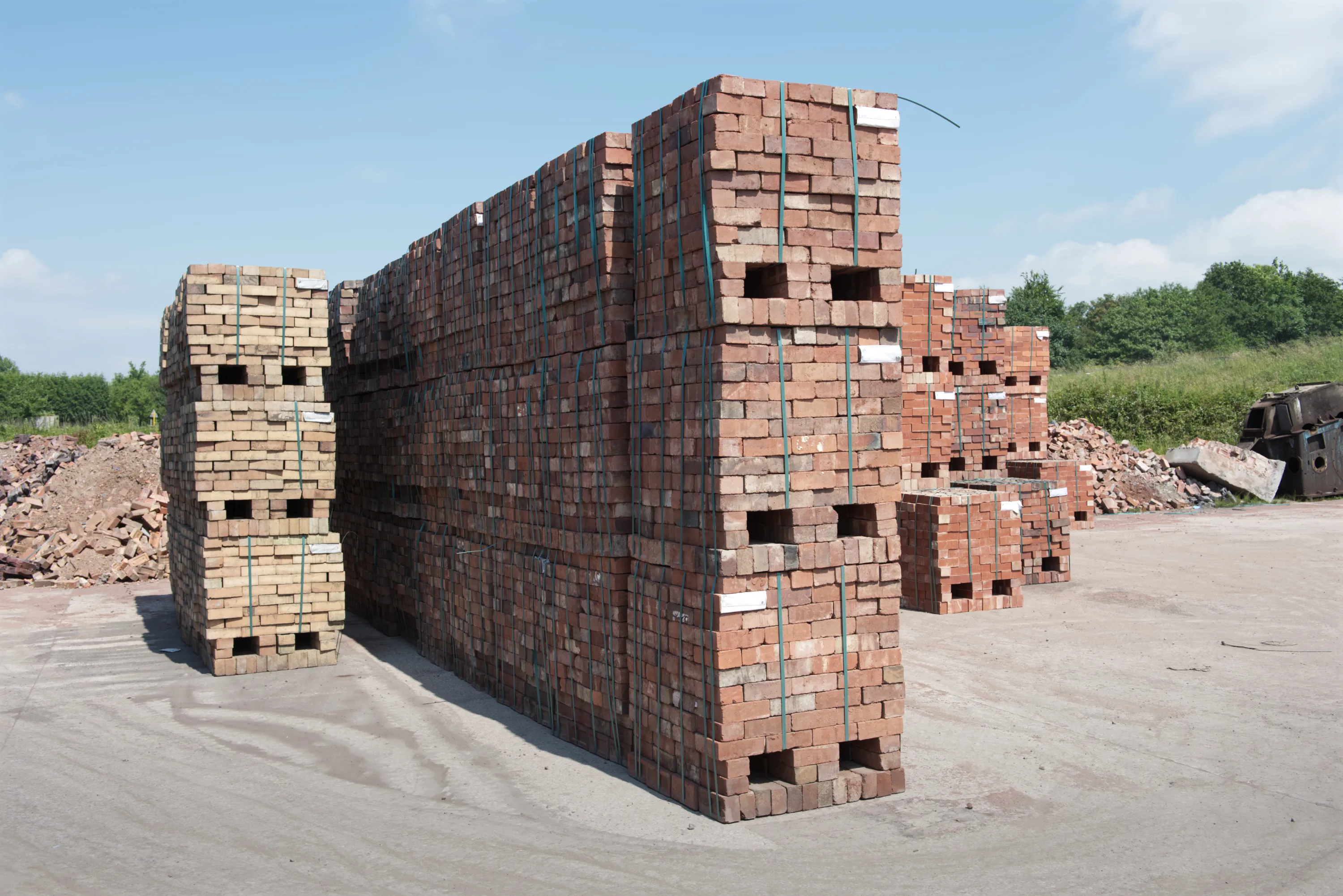 Pallets of fired red bricks stacked on a concrete yard, with green trees and a blue sky behind