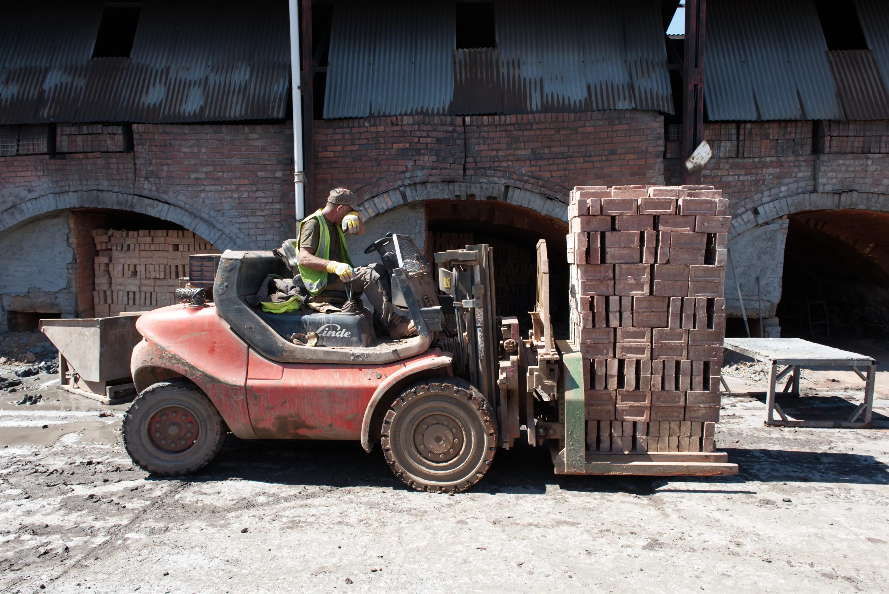 A worker in a hi-vis vest operating a red forklift carrying a stack of bricks outside a kiln arch