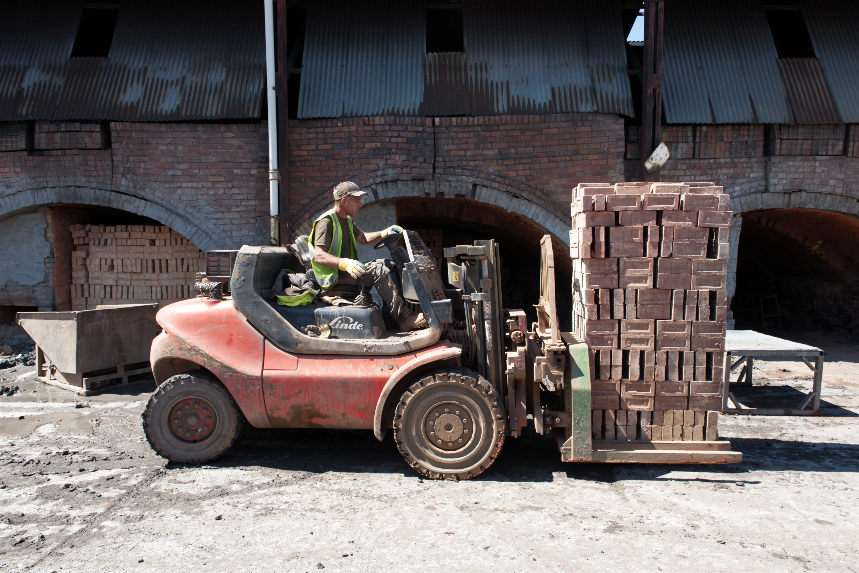 A worker in a hi-vis vest driving a red forklift with a load of bricks beside a brick kiln arch