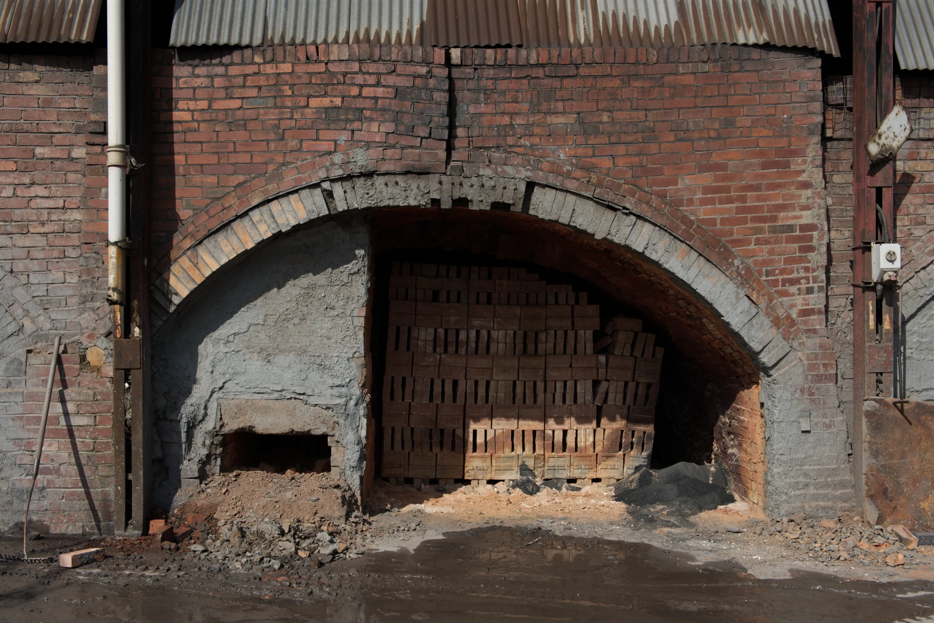 Front view of a kiln arch partially bricked up, with raw bricks stacked inside and rubble at the base