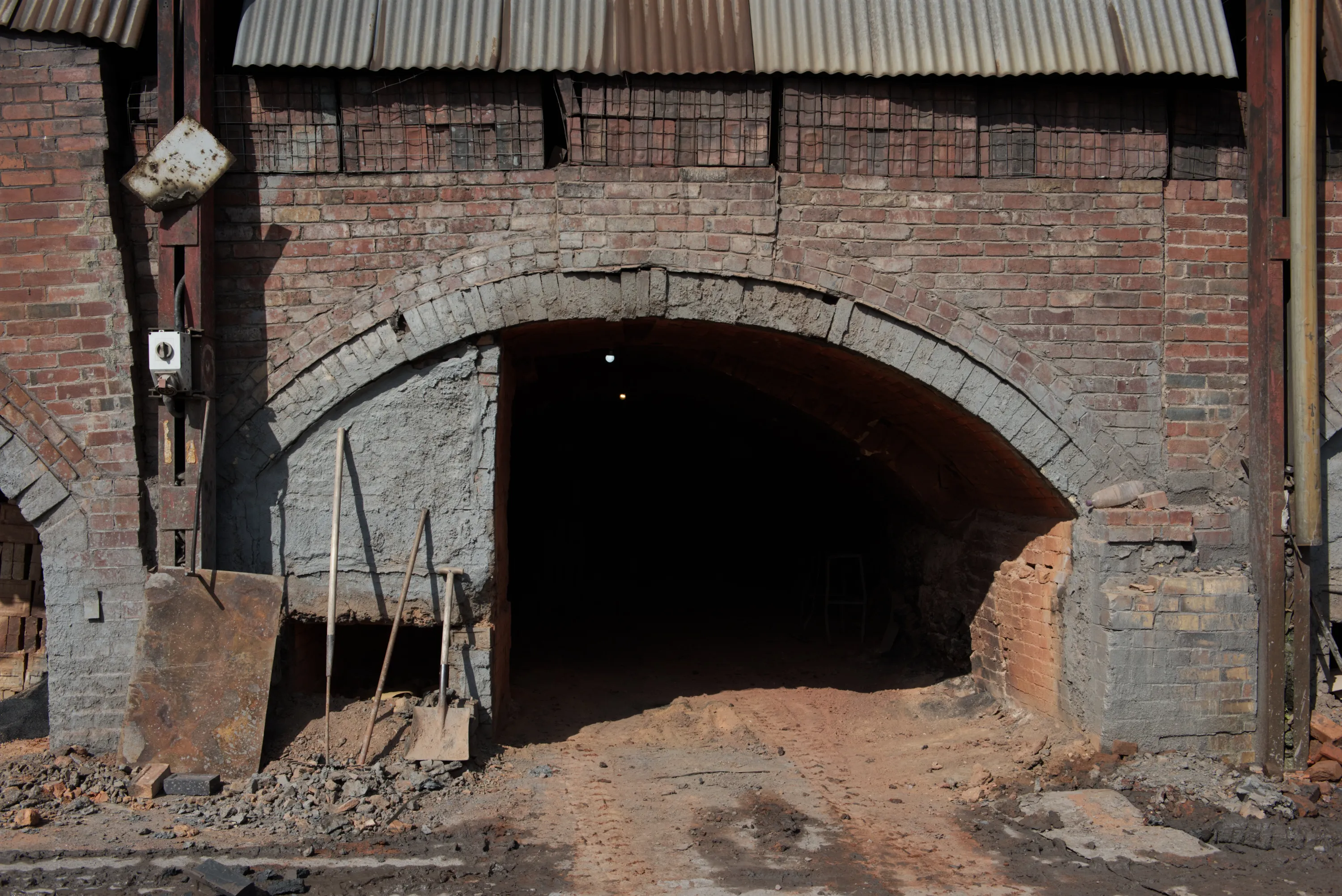 An open kiln arch in full darkness, tools leaning against the wall, a single bulb glowing inside