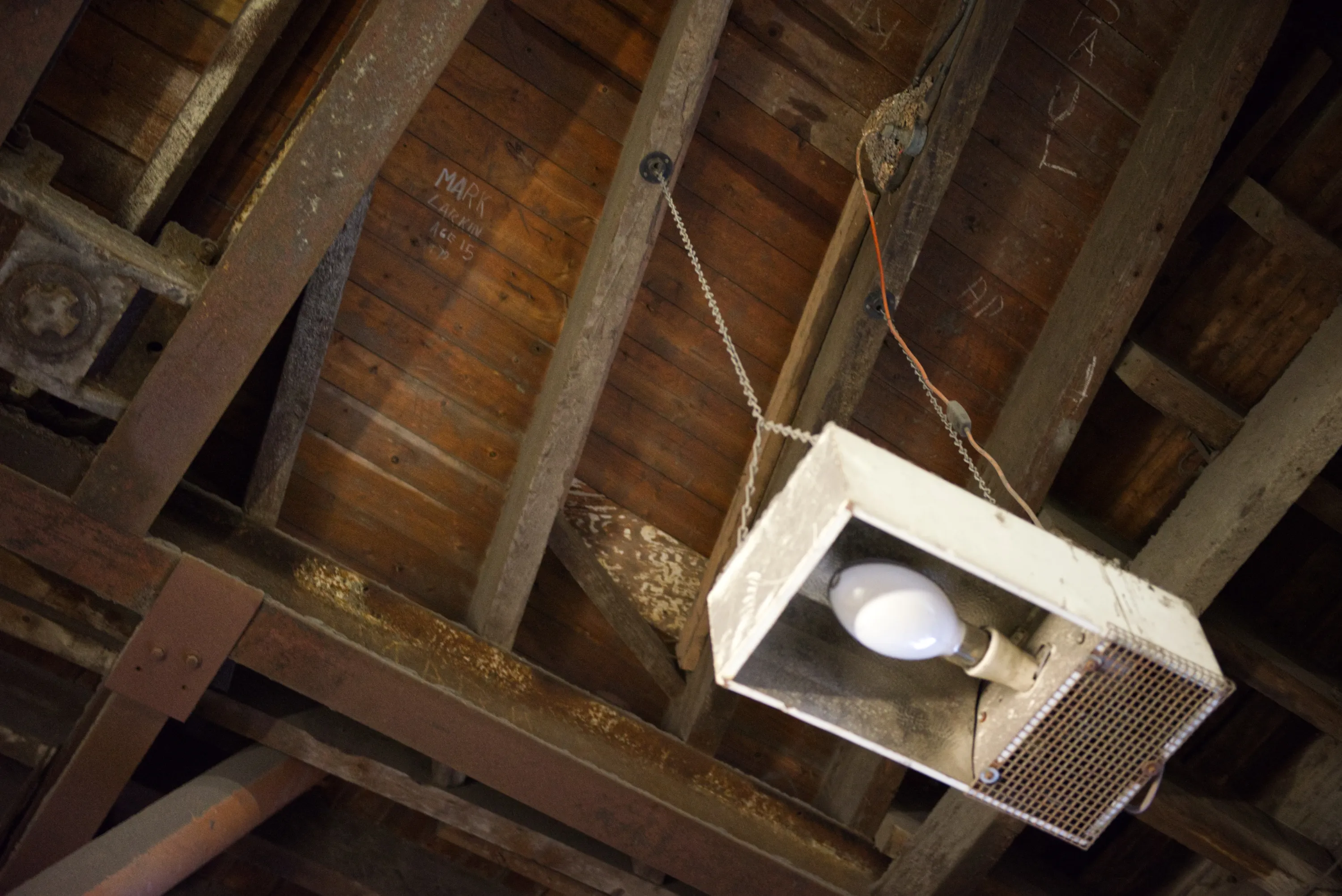 A dusty industrial light fitting hanging from old wooden roof timbers, names written on the wood behind