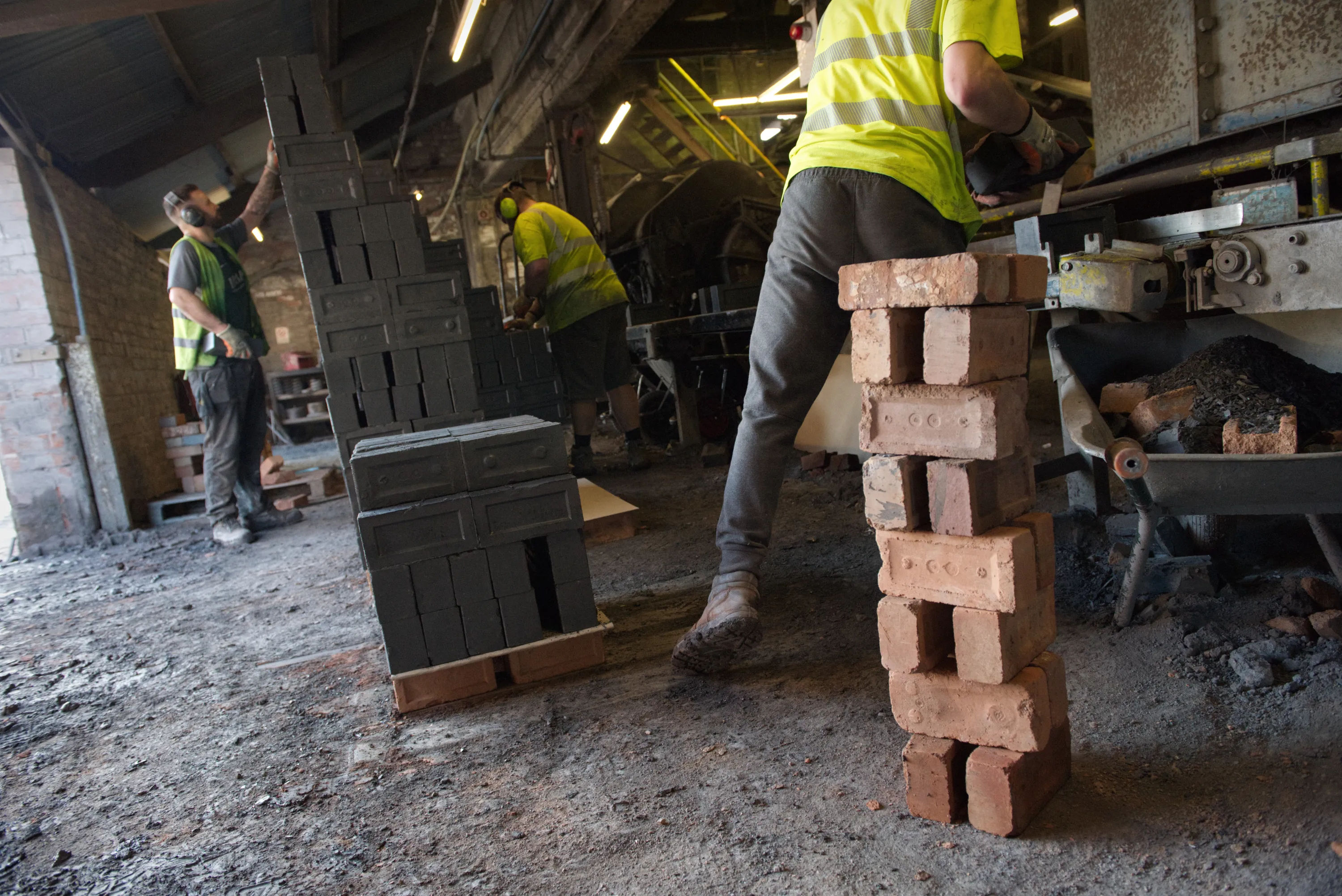 Three workers in hi-vis vests handling bricks on the floor of a large brick shed
