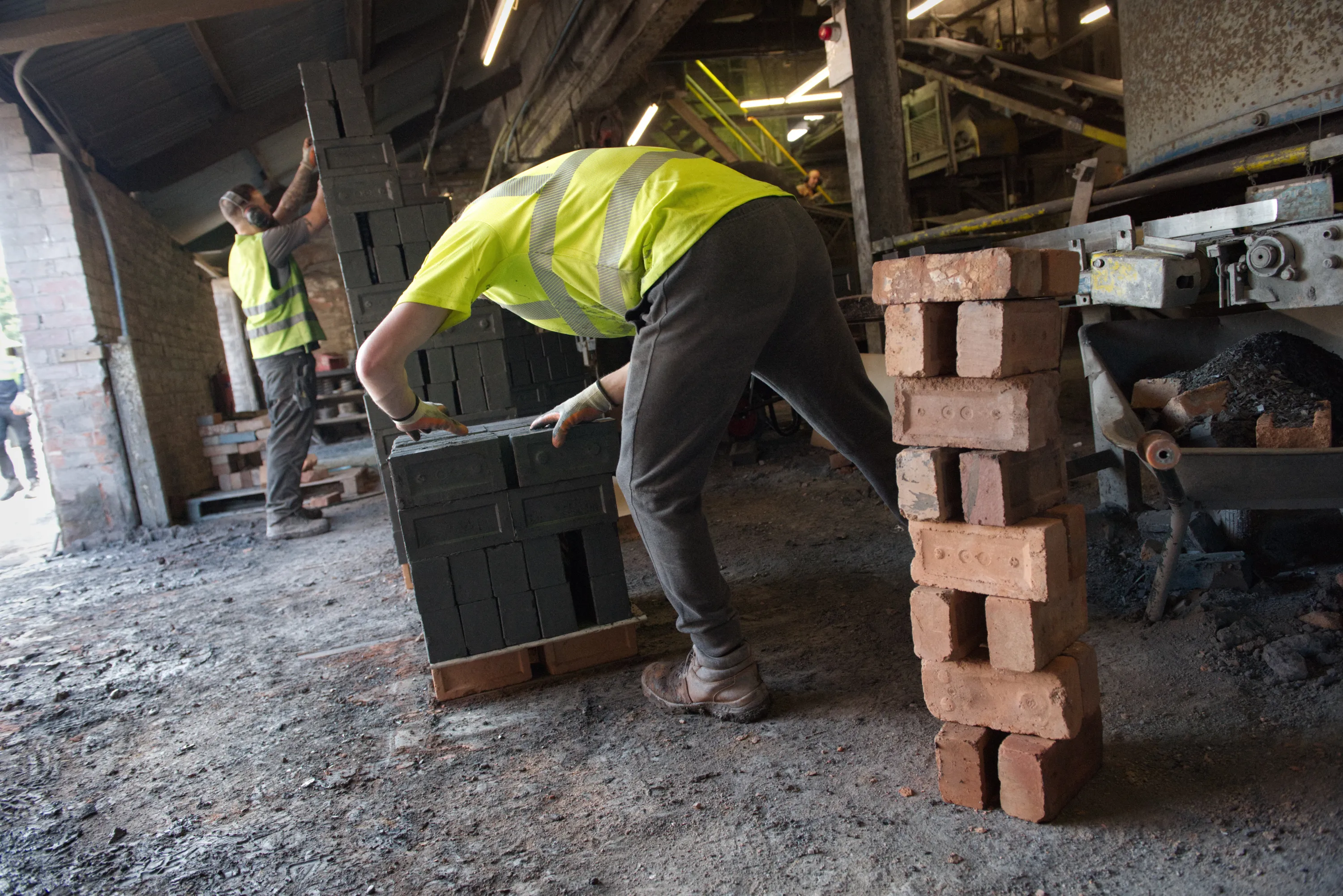Two workers in hi-vis vests stacking bricks on pallets inside an industrial shed