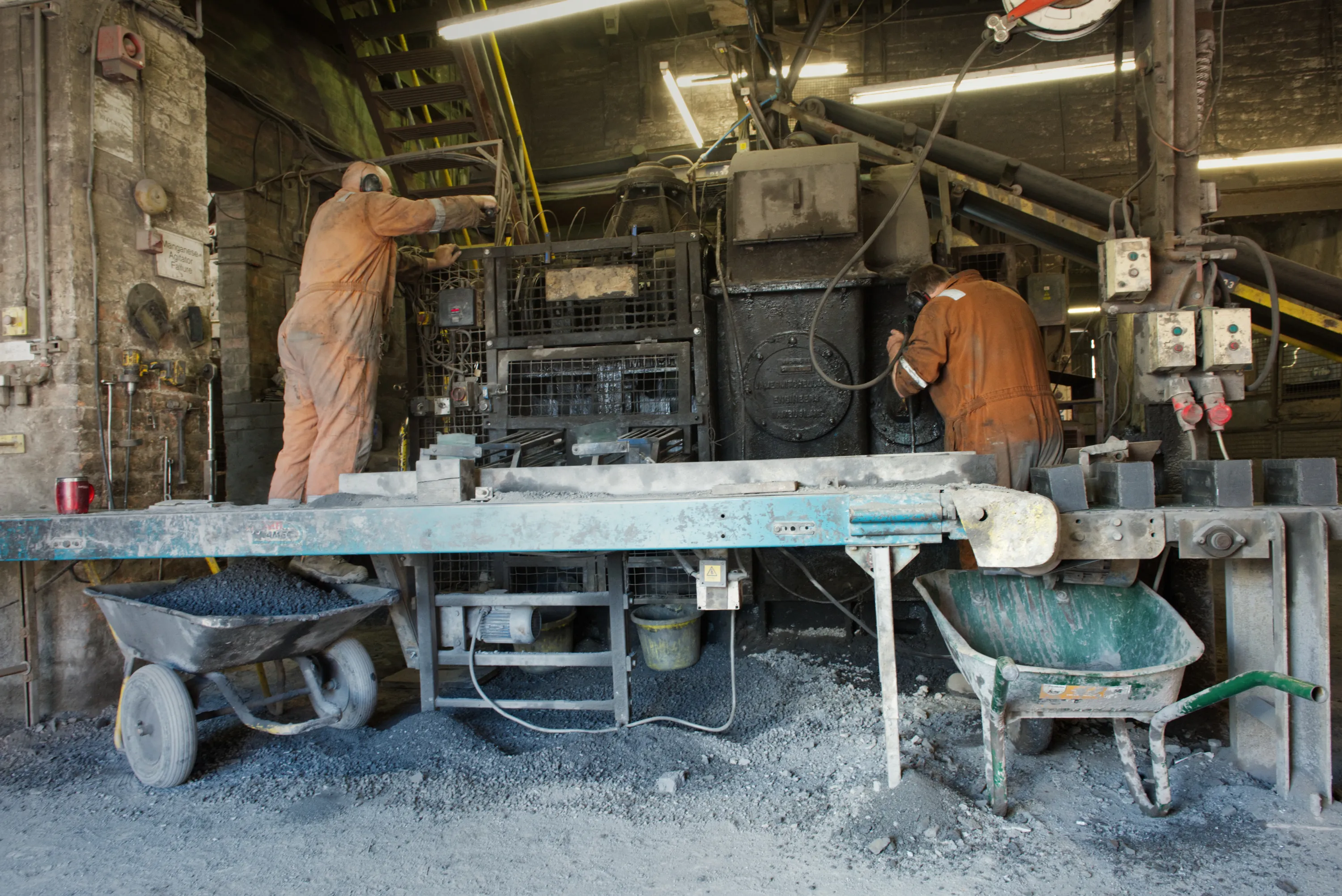 Two workers in overalls servicing heavy machinery beside a conveyor belt, wheelbarrows below
