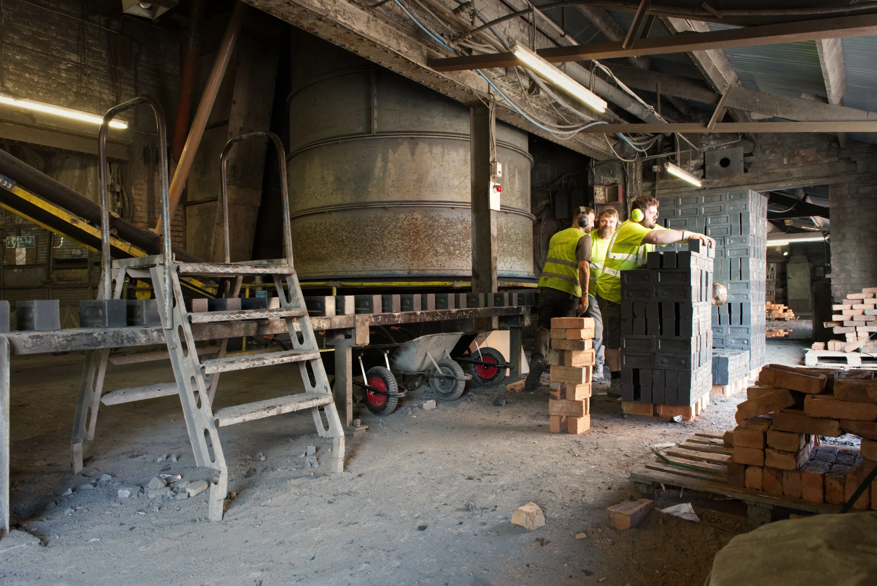 Workers in hi-vis vests moving bricks beside industrial equipment inside the works