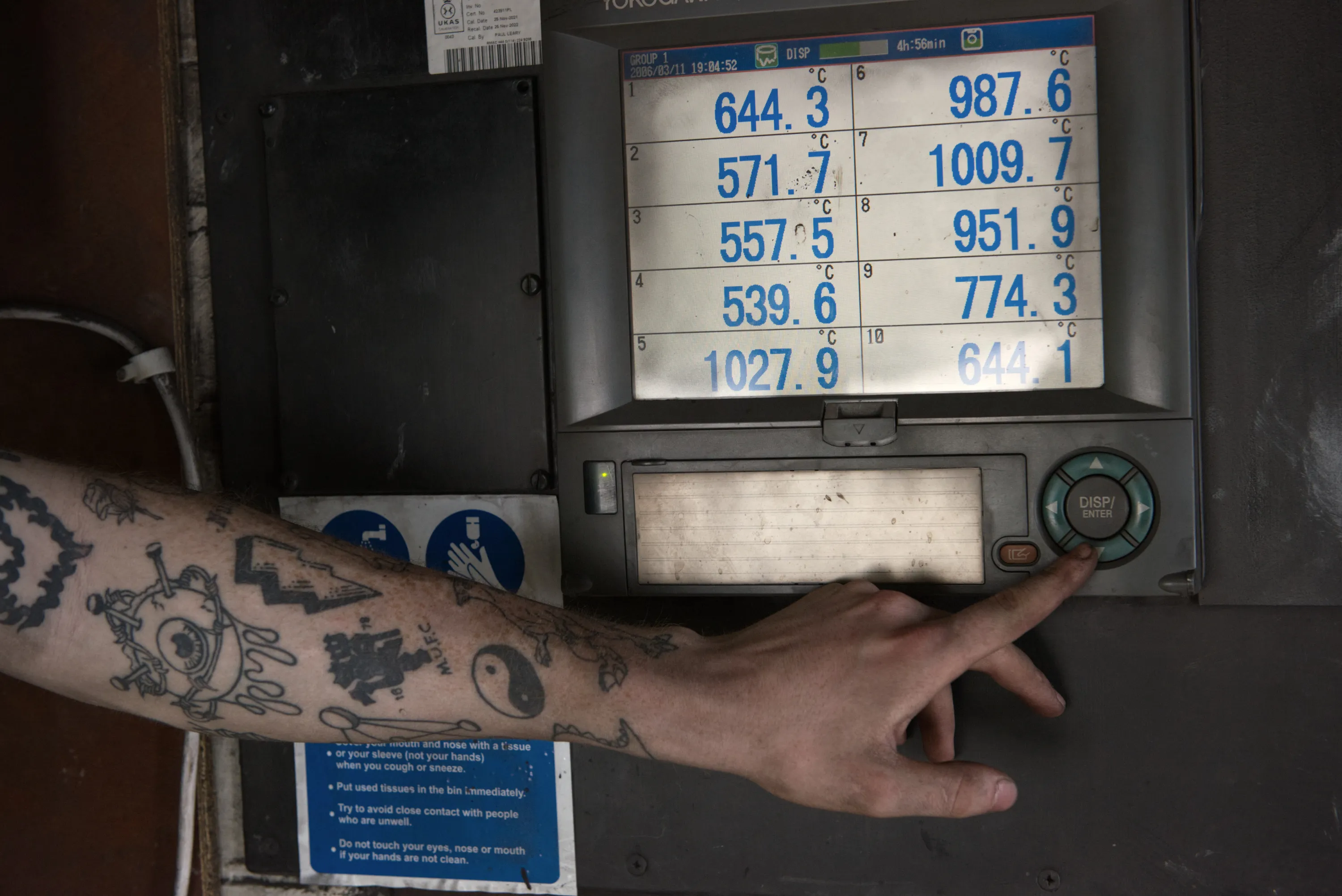 A tattooed hand pointing at a kiln temperature controller displaying readings between 539 and 1027 degrees