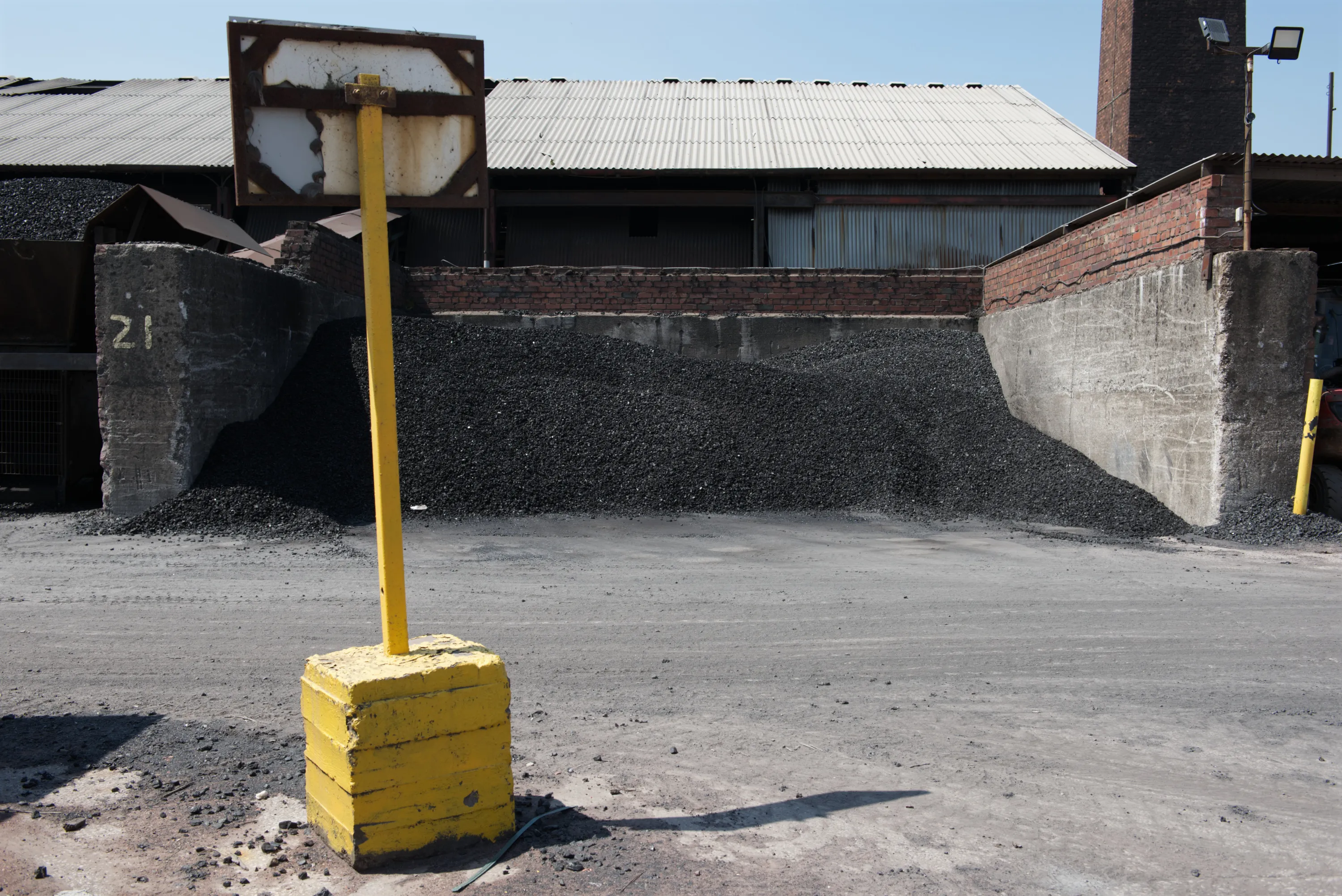 A yellow bollard in front of a large coal stockpile against corrugated buildings and a brick chimney