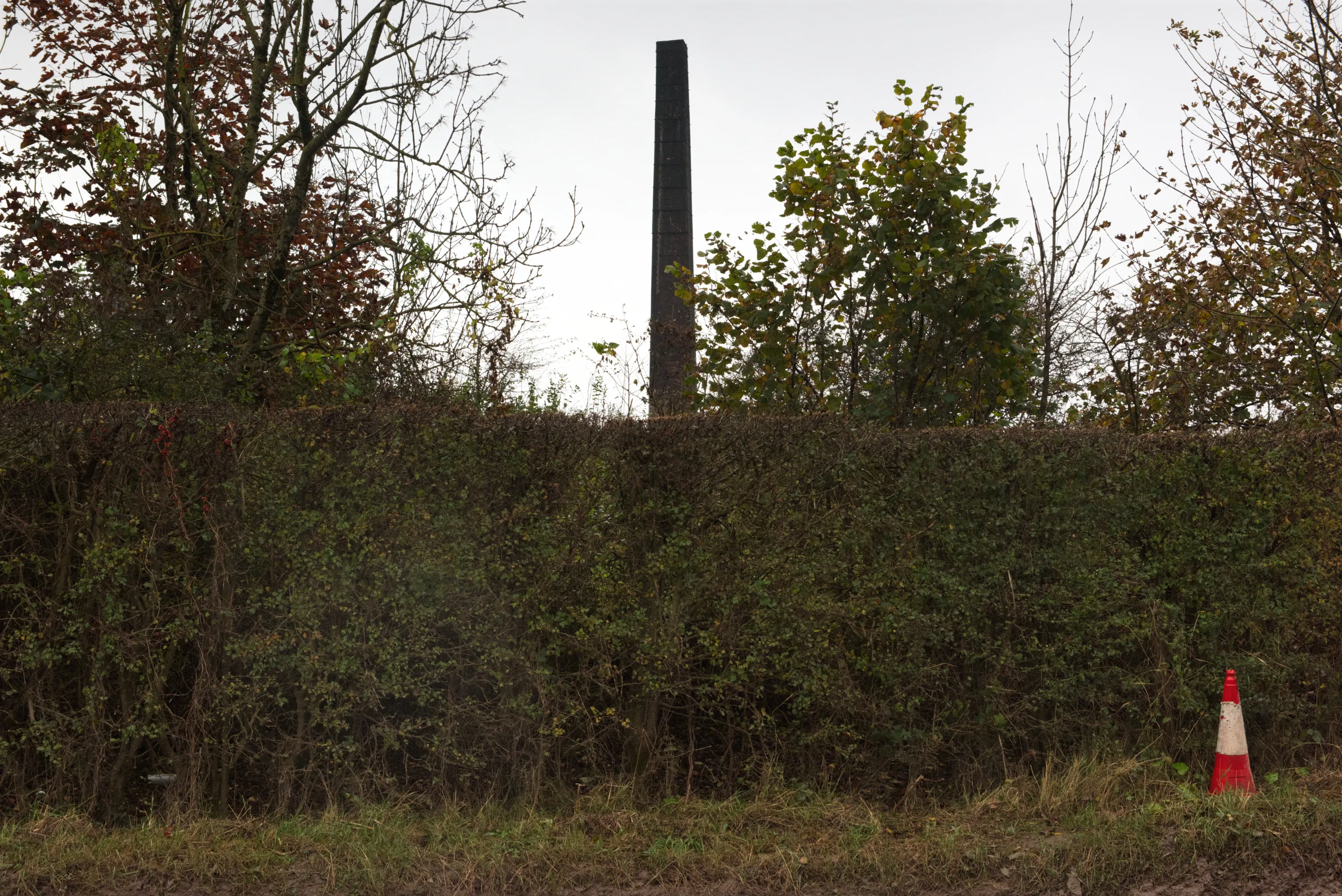 The brickworks chimney rising above a hedgerow, a traffic cone in the foreground on an overcast day