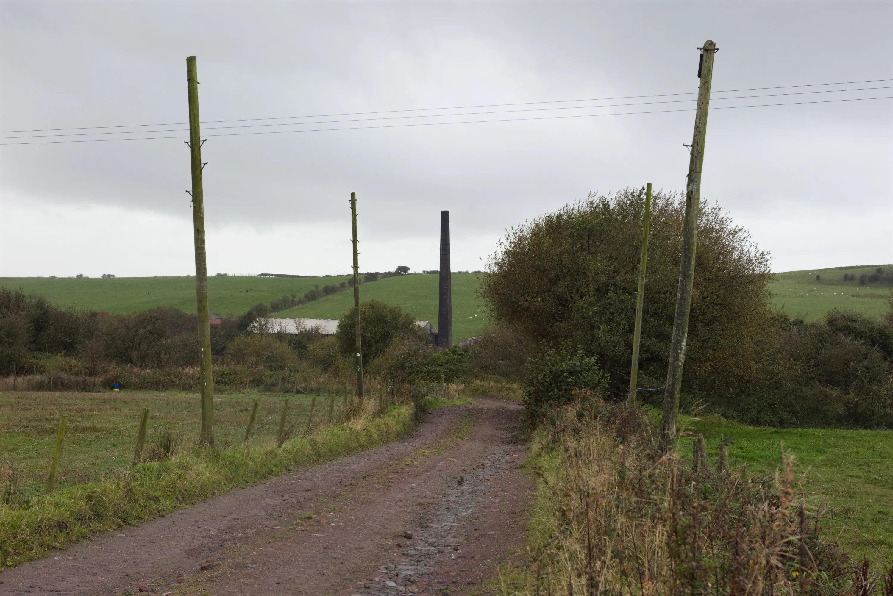 A rural track lined with telegraph poles and scrub, the brickworks chimney rising in the middle distance