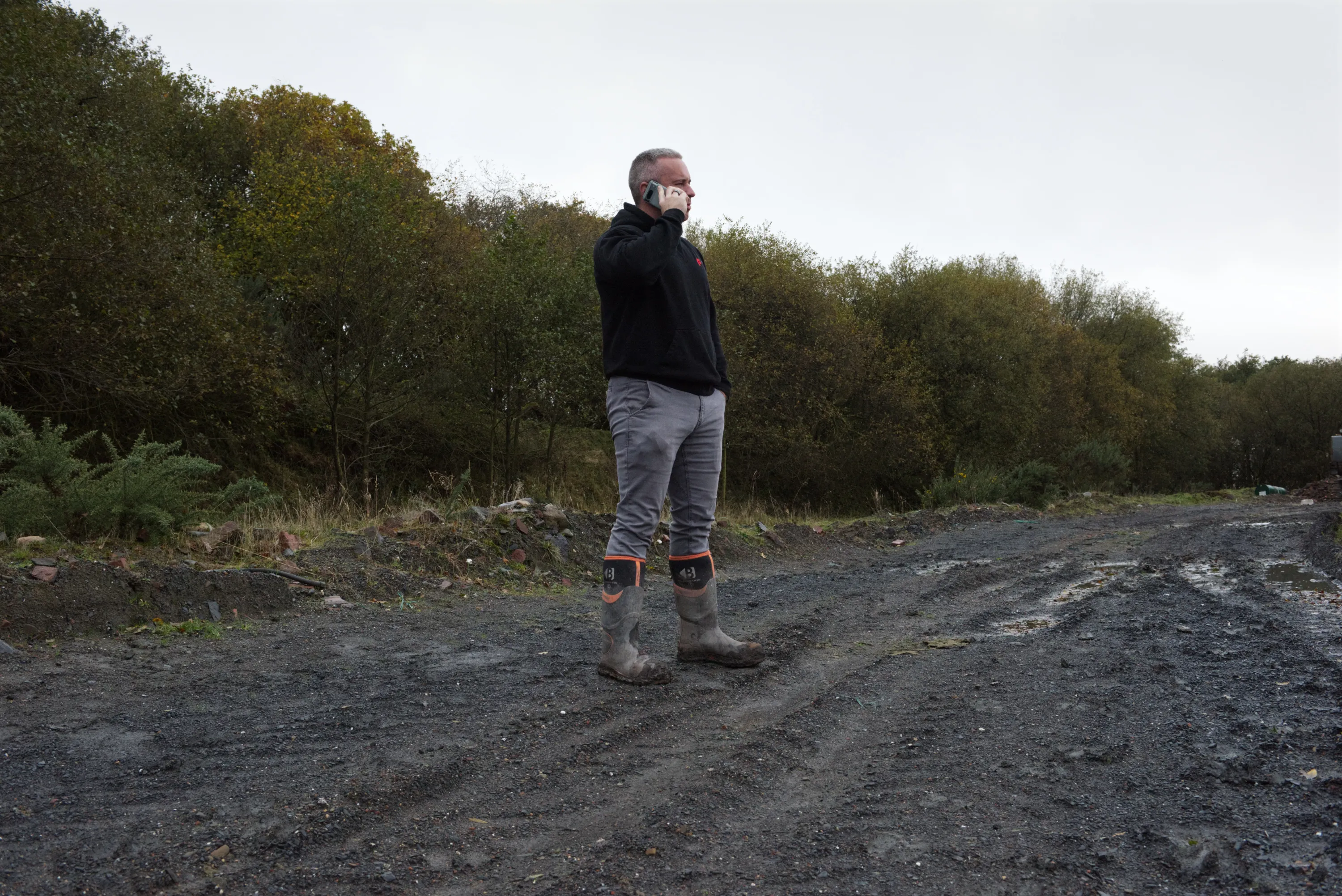 A man in wellington boots talking on a mobile phone in a muddy quarry, trees behind him
