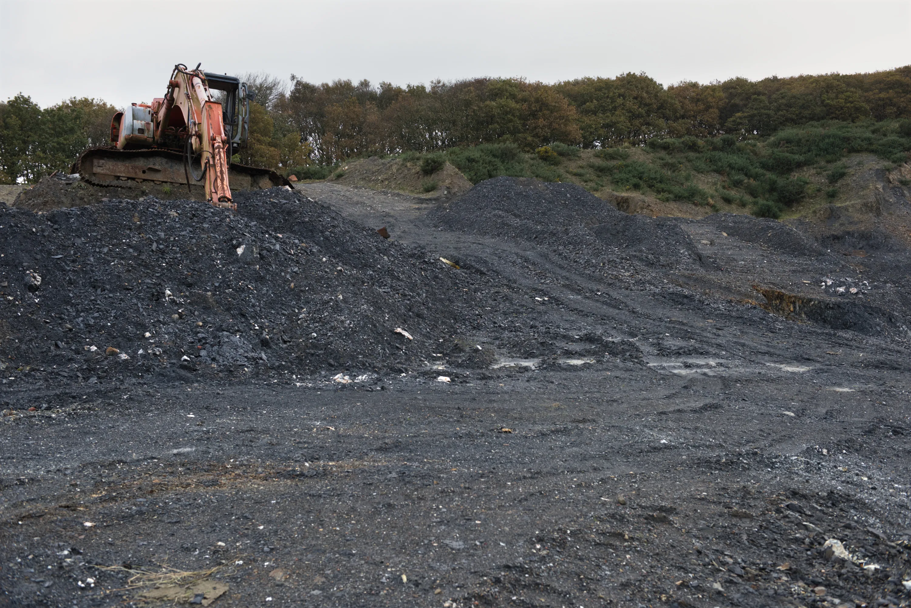 An excavator perched on a mound of dark shale spoil in an open quarry, trees on the ridge behind