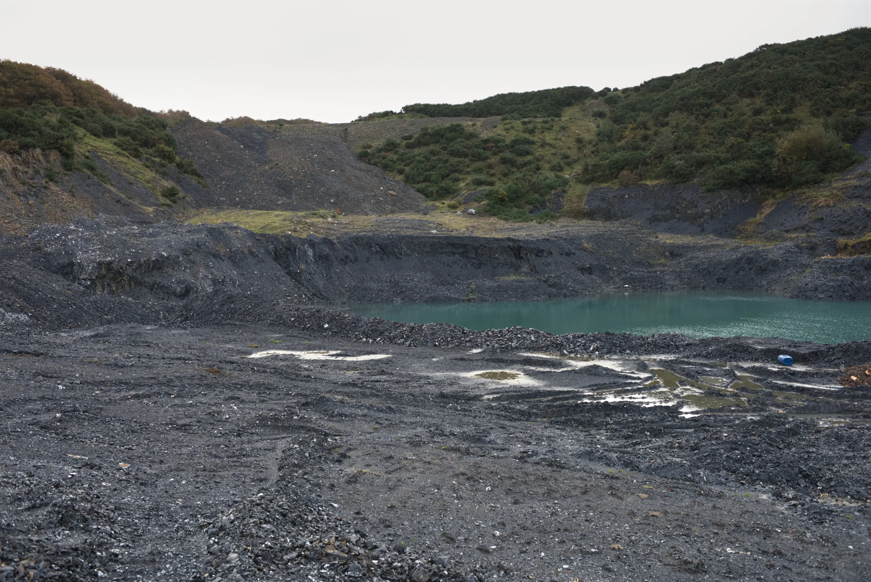 A shale quarry with dark spoil heaps, a turquoise pool of standing water at its floor, overcast sky