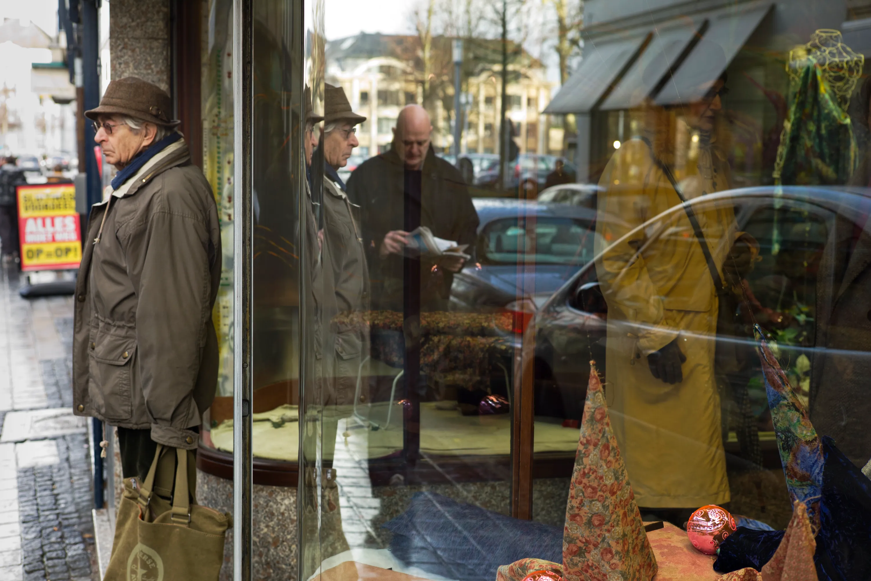 Older man in hat and jacket standing outside a shop window, reflections of street and mannequins