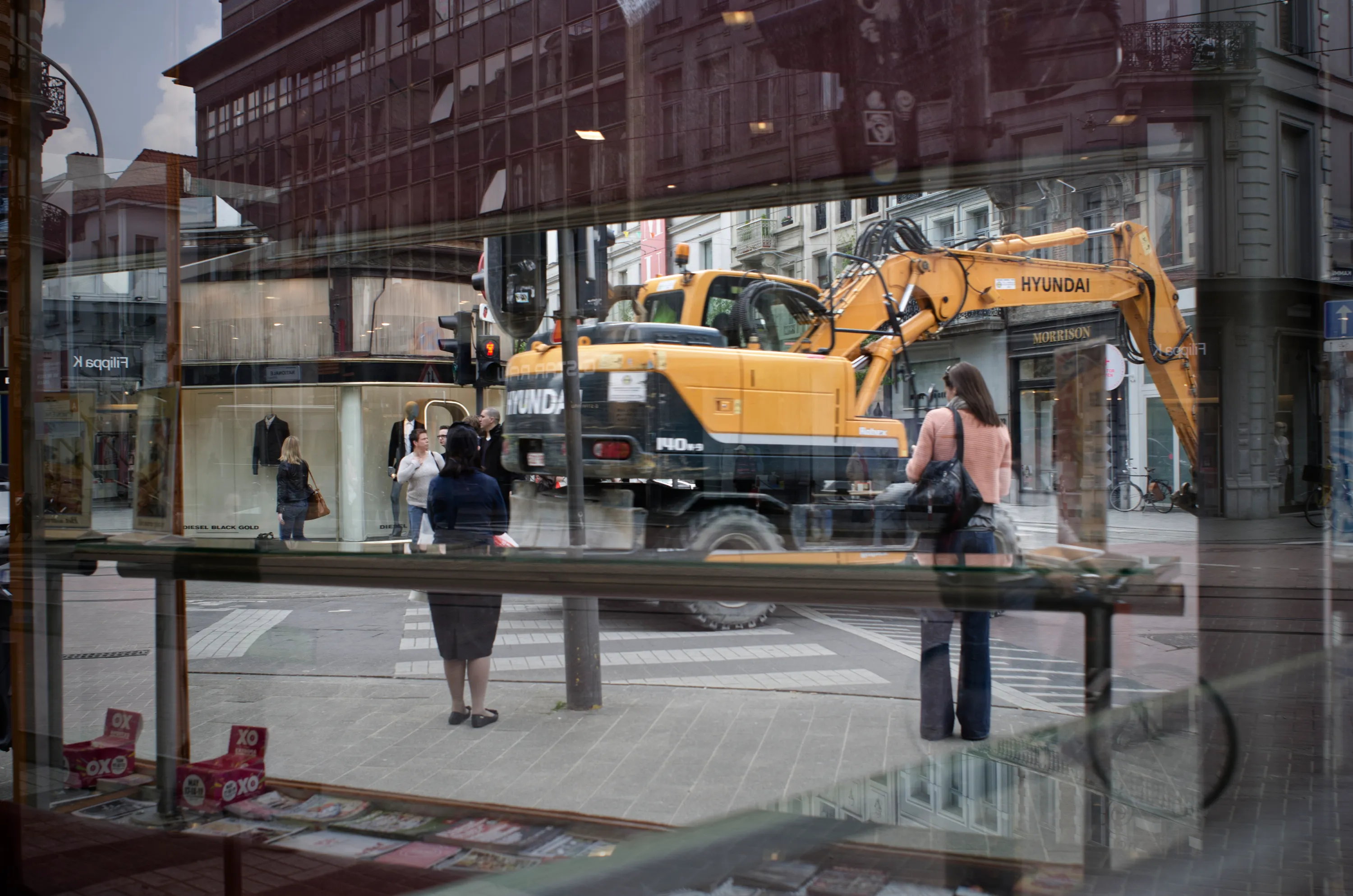 City street seen through glass, yellow excavator and pedestrians reflected in the pane