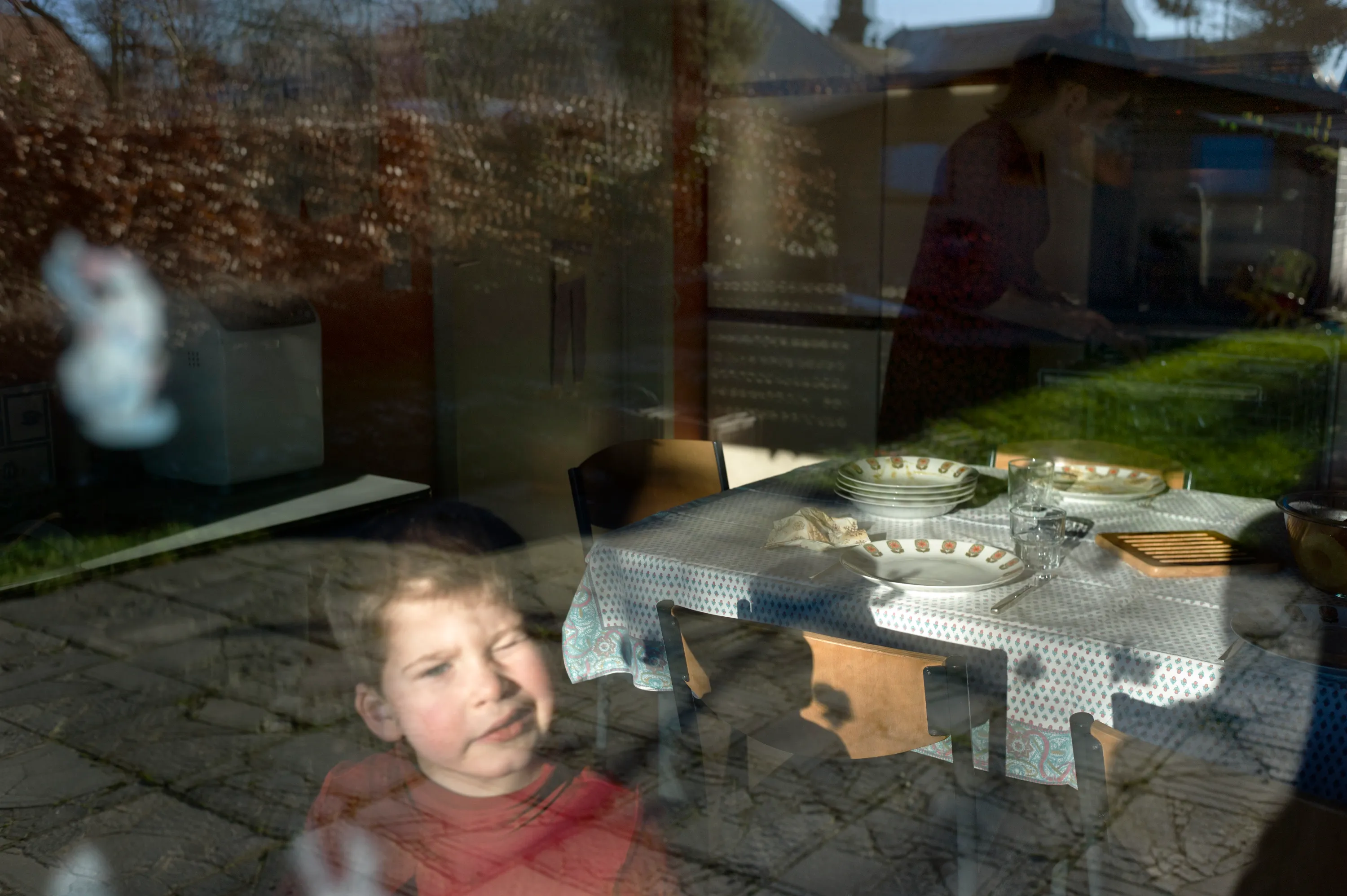 Young child at a window, dining table with stacked plates visible inside and garden beyond