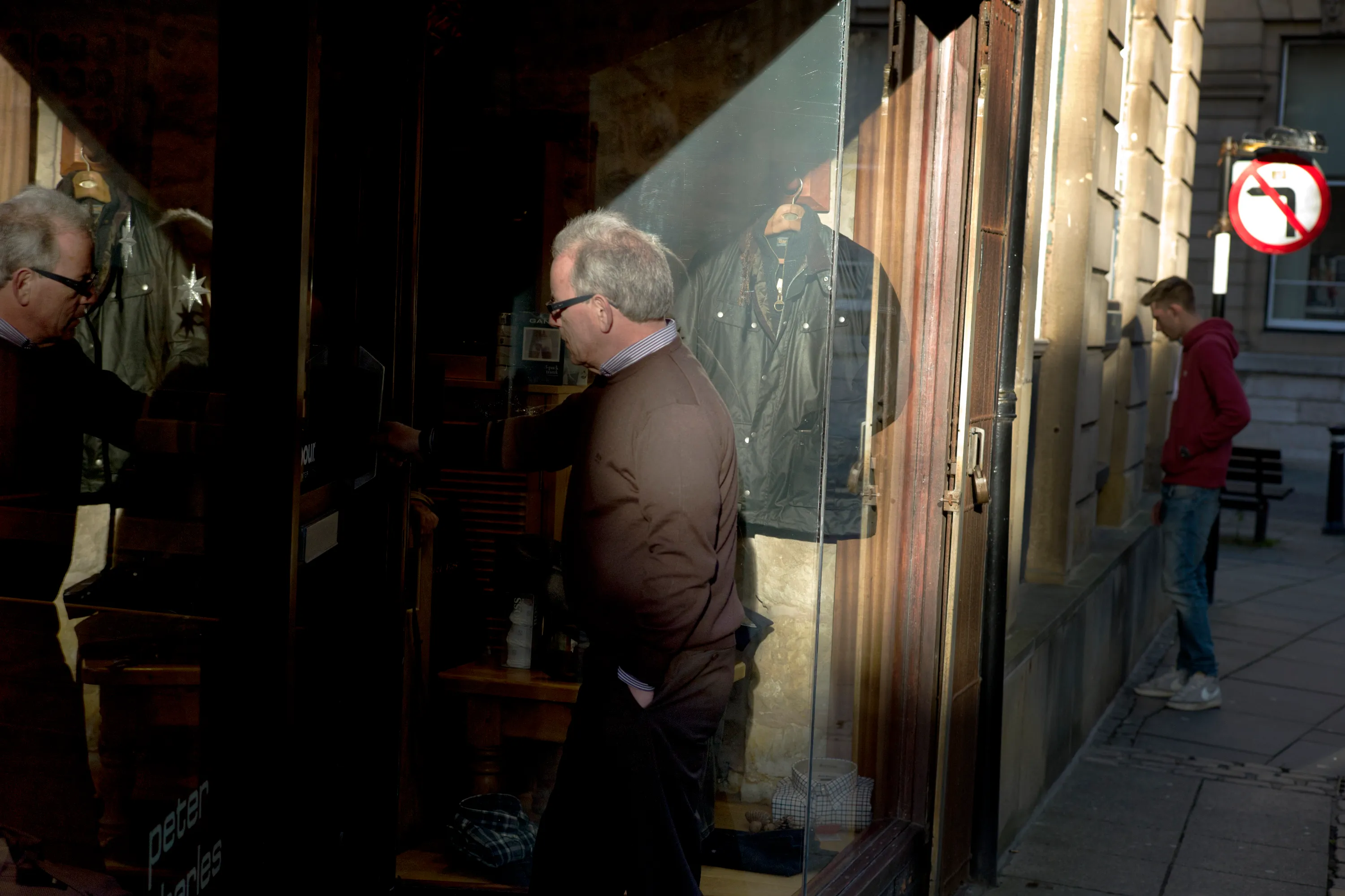 Man in brown jacket peering into a shop window, leather jacket on display inside