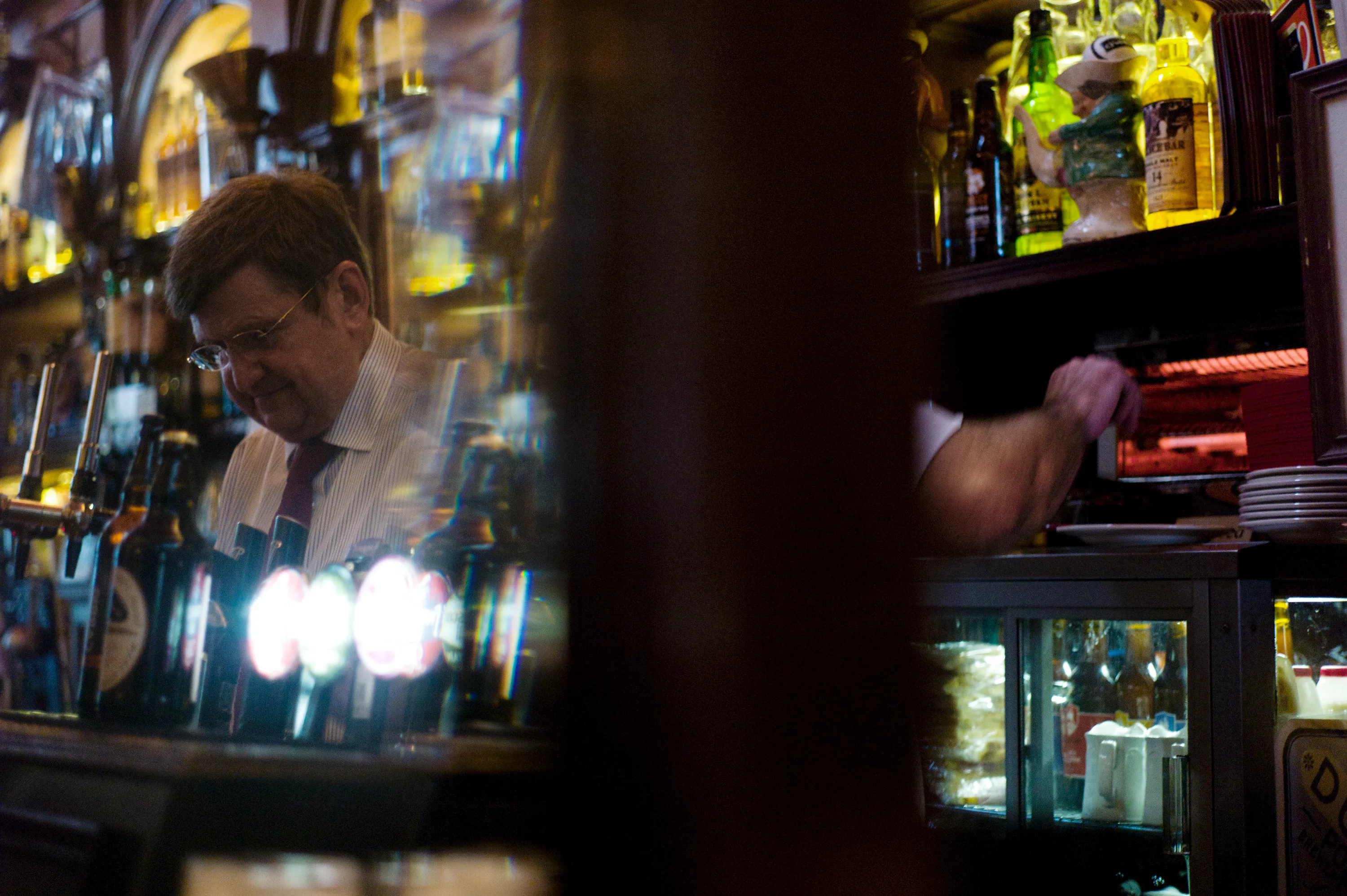 Barman behind a pub bar, tap handles and bottles of spirits on shelves behind him