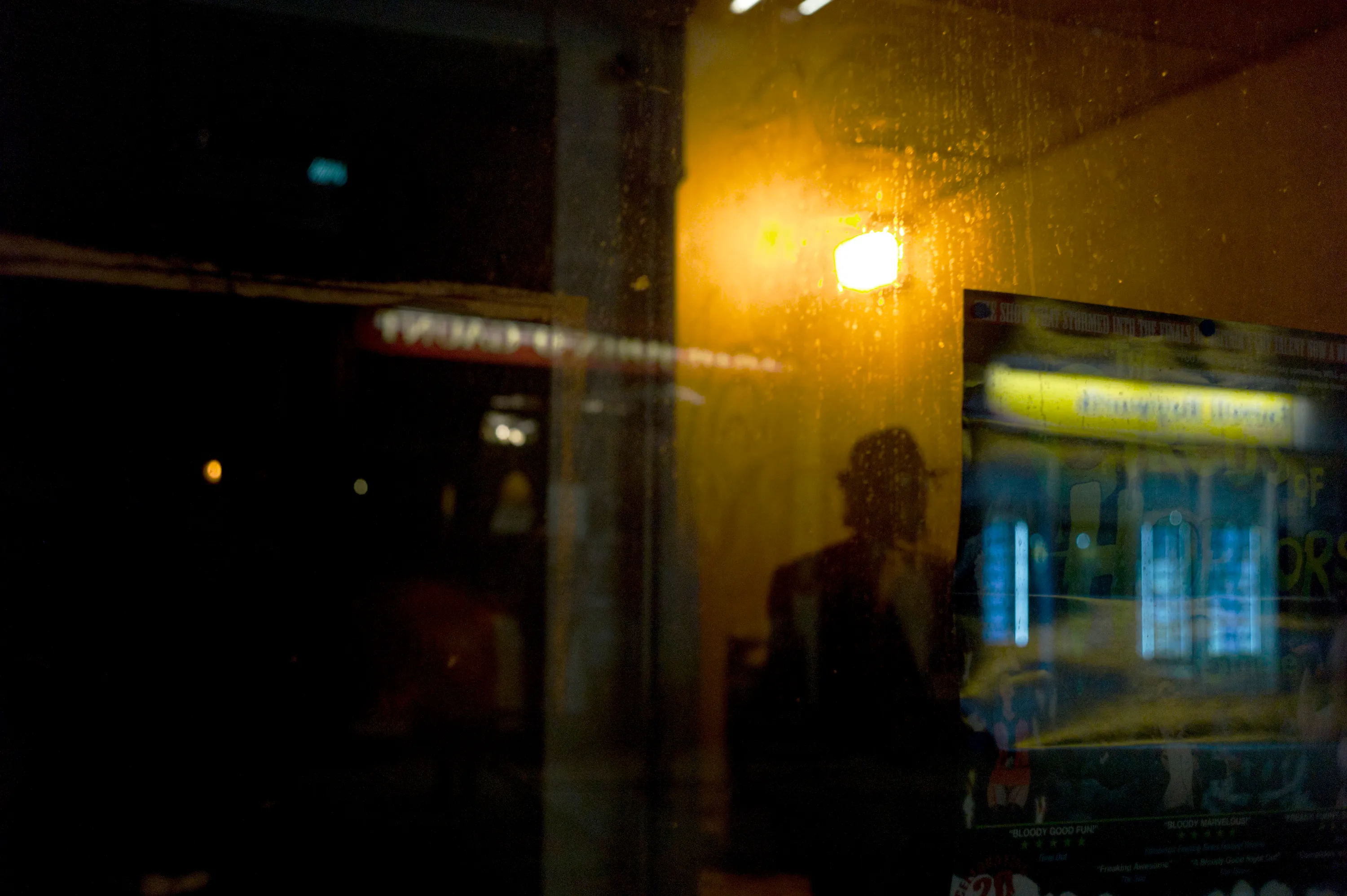 Silhouette reflected in a rain-speckled window at night, warm lamp glow behind