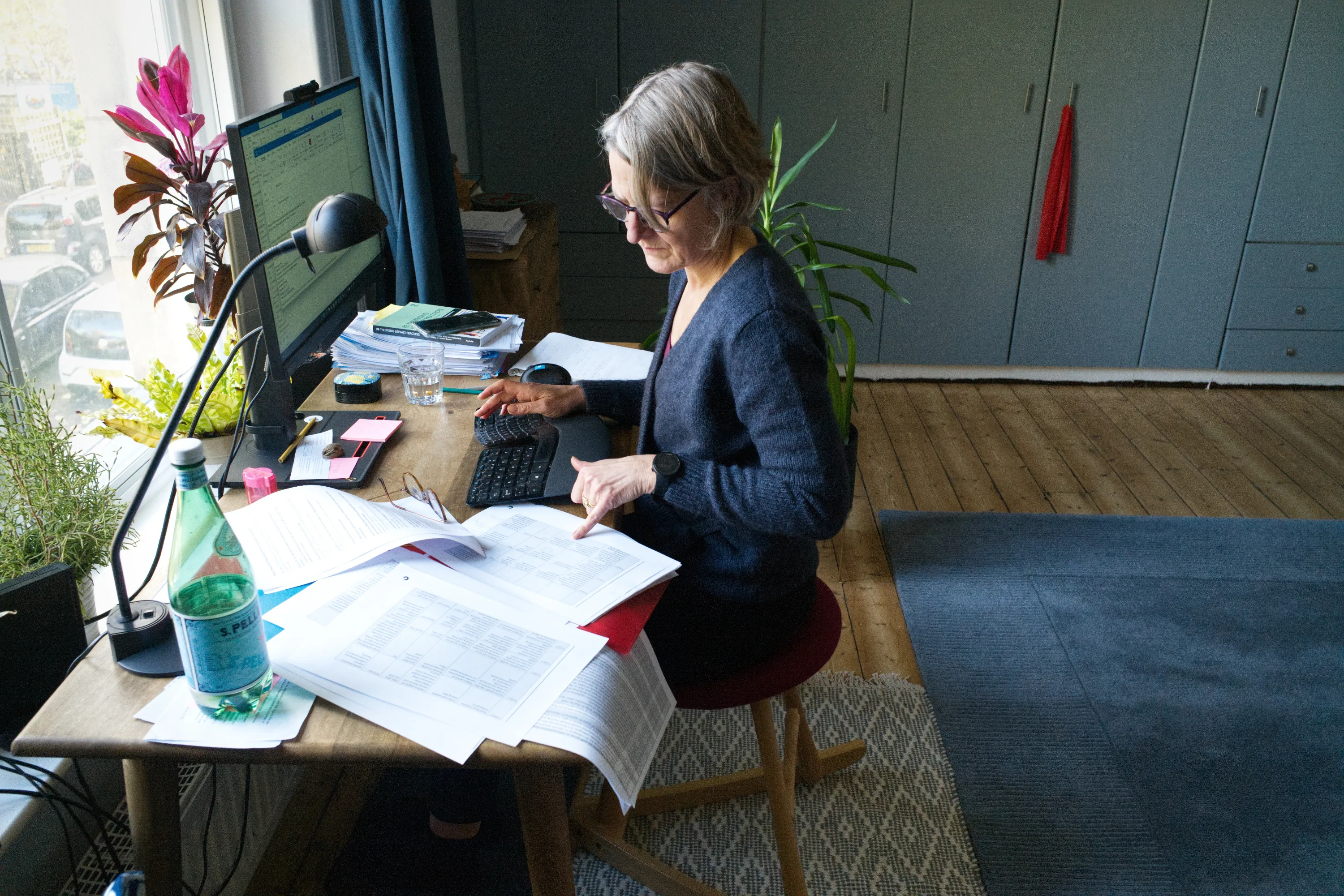 Woman at a desk reviewing printed documents beside a large monitor and window