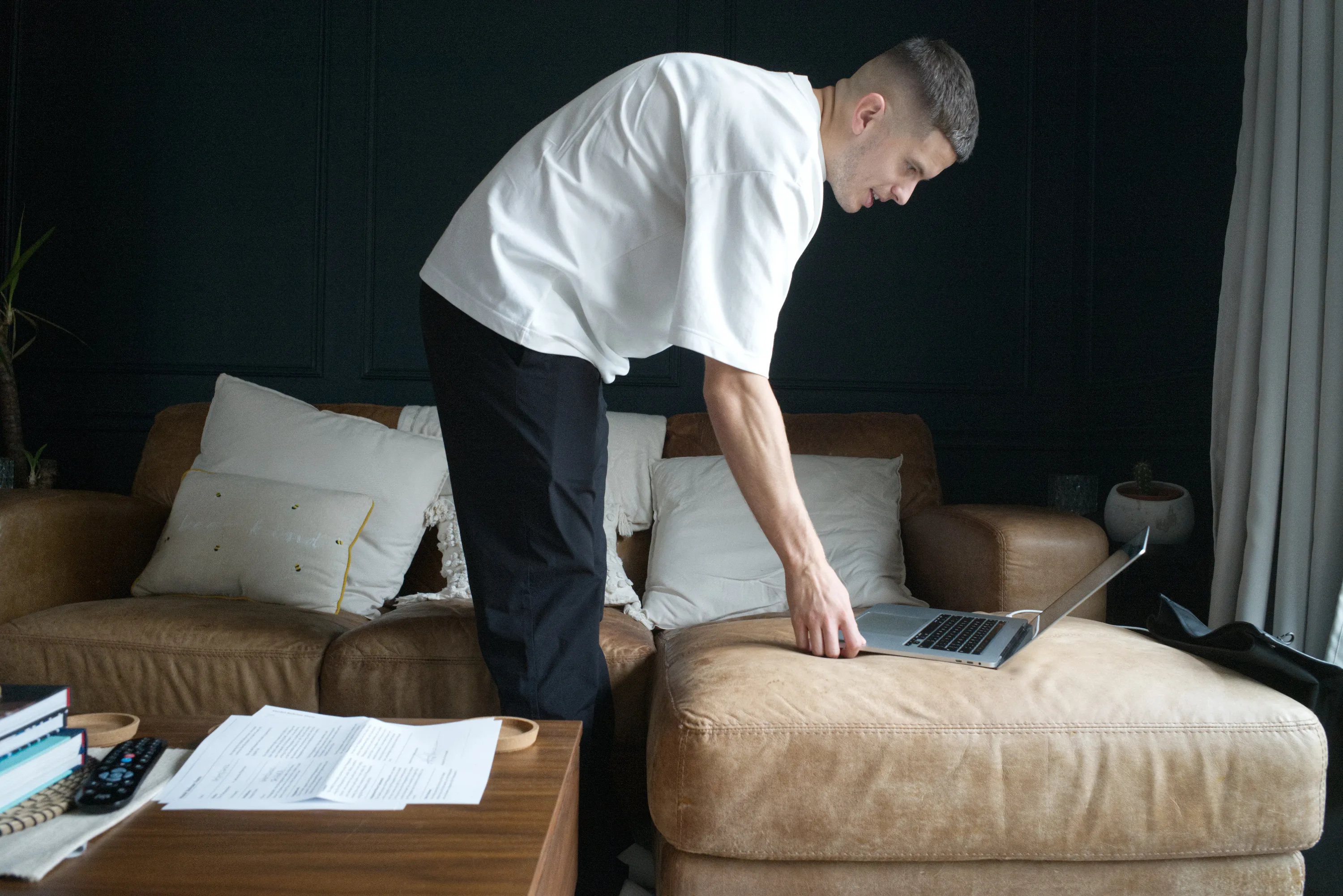 Man leaning over a sofa arm to use a laptop in a dark living room