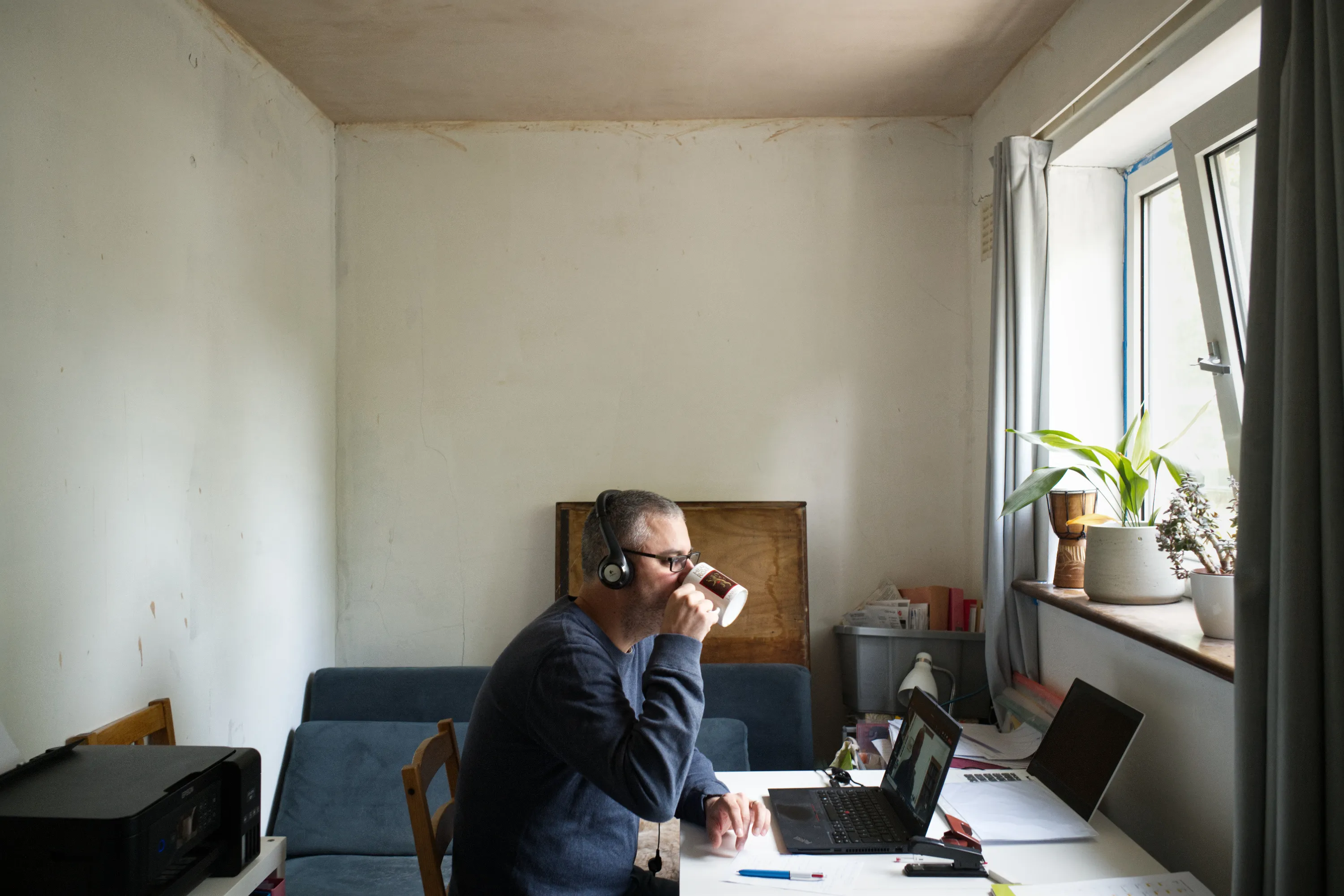Person wearing headphones drinking from a mug at a laptop by a window