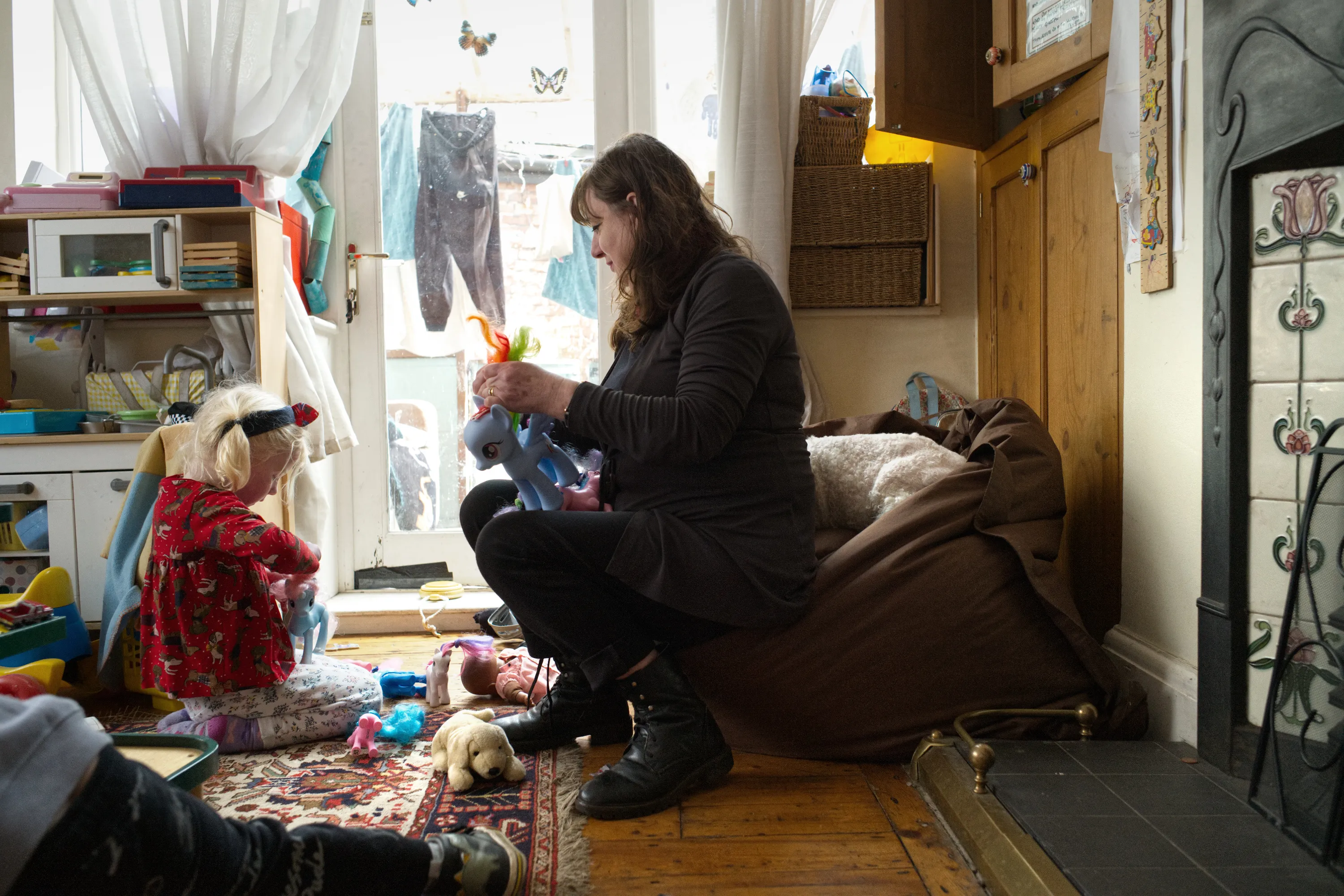 Woman kneeling on a floor holding a toy beside a young child amid scattered toys