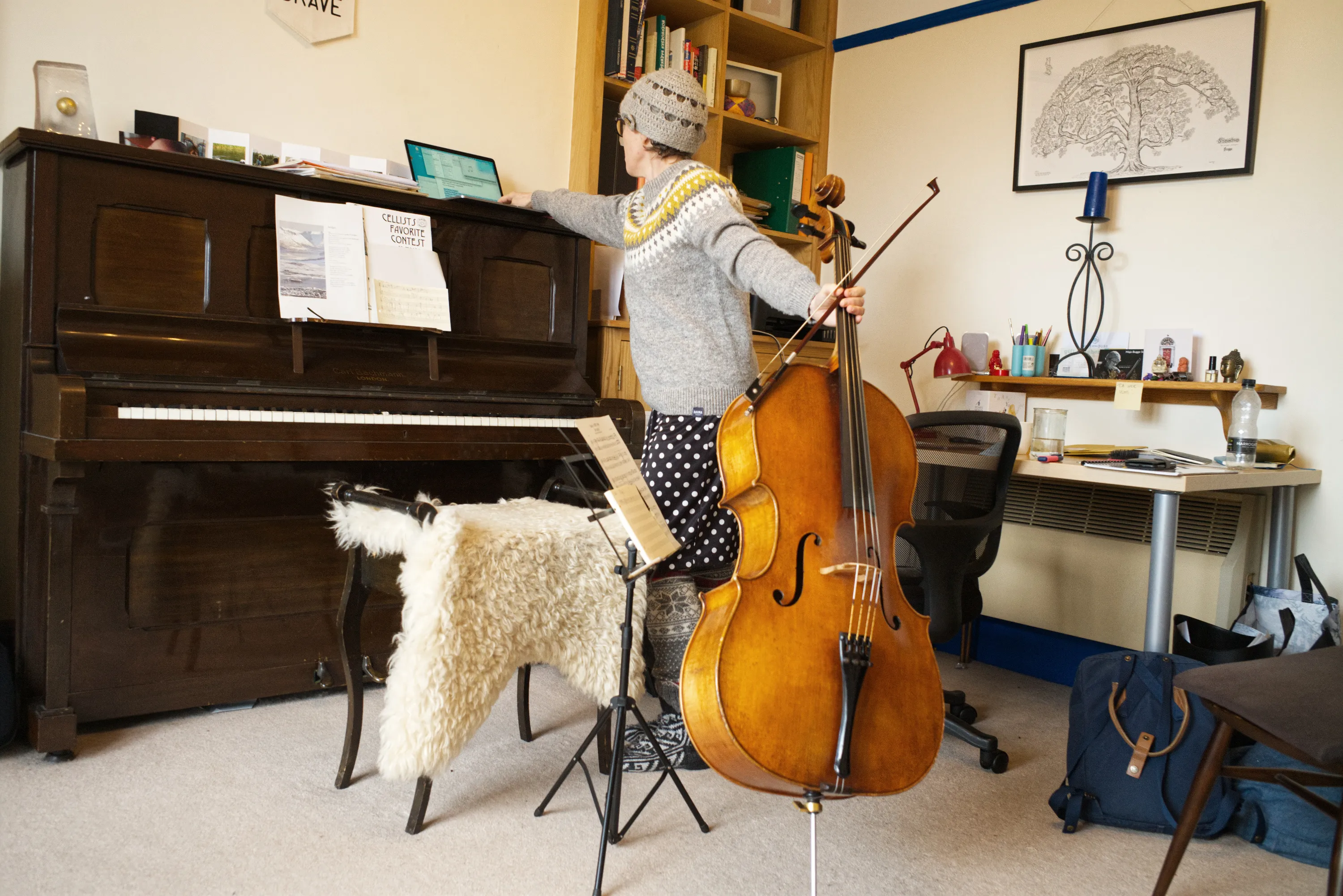 Person standing playing cello in a room with an upright piano and bookshelf