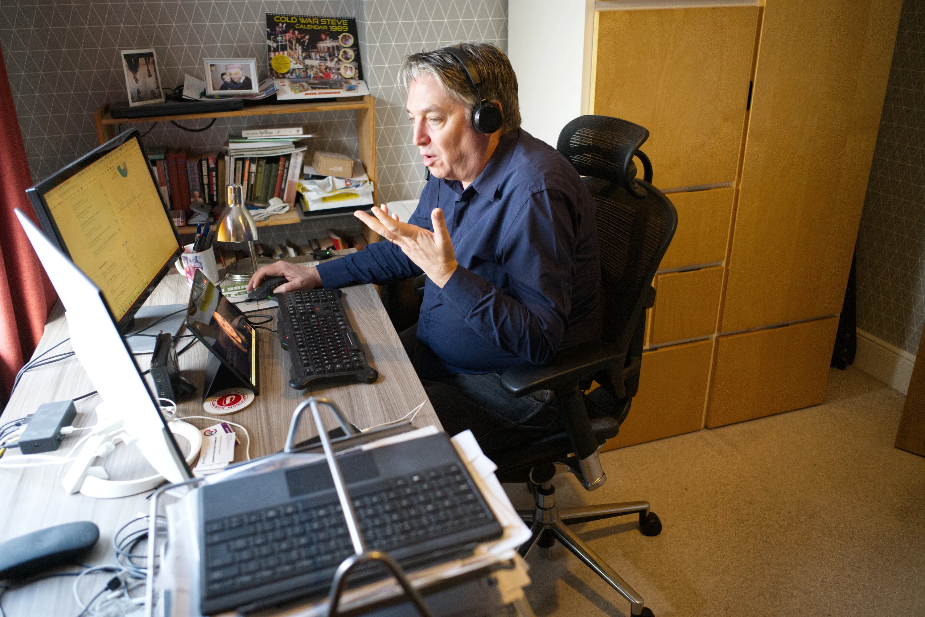 Man wearing headphones gesturing at dual monitors on a cluttered desk
