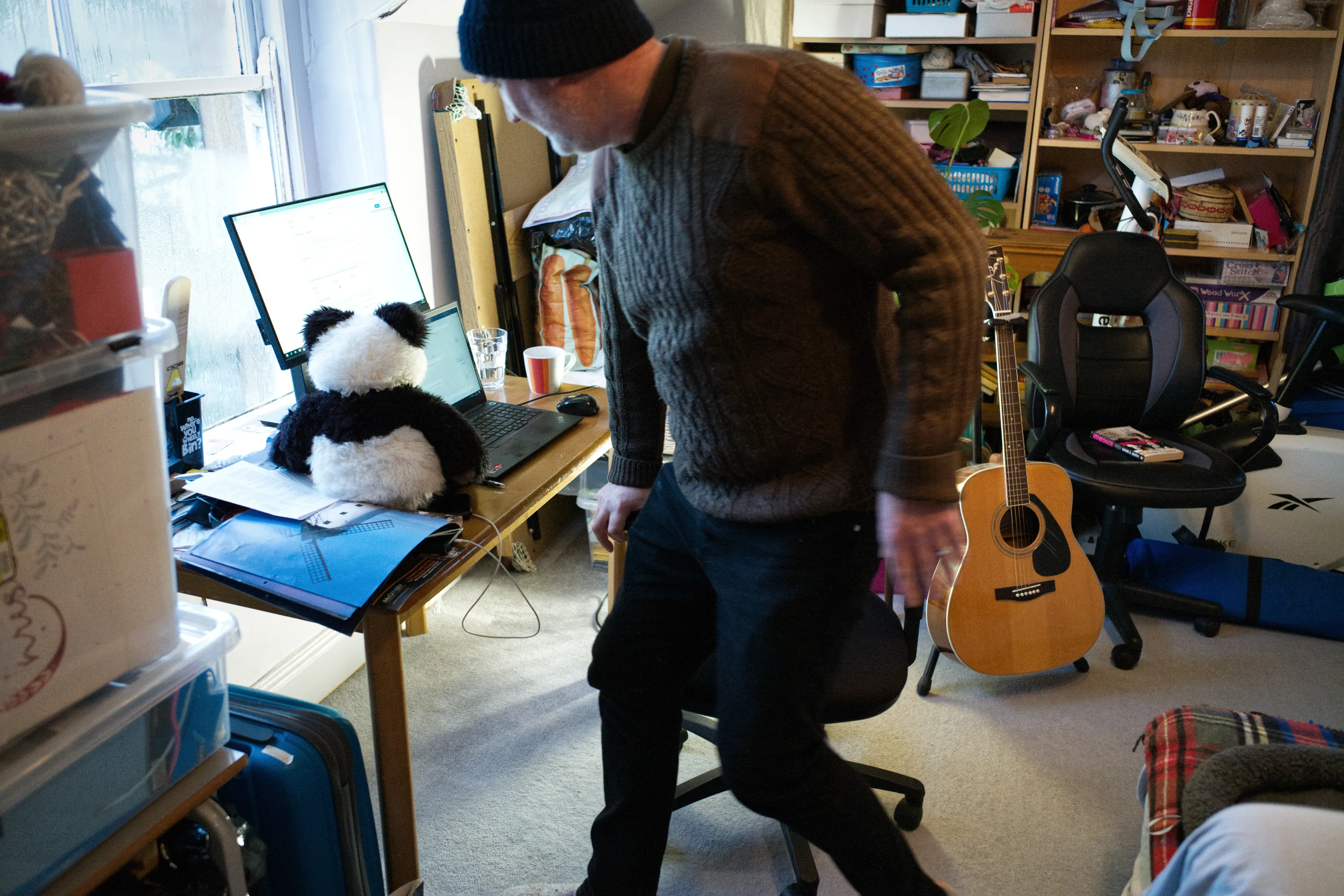 Man in a beanie moving in a cluttered room with a laptop, guitar leaning against the wall