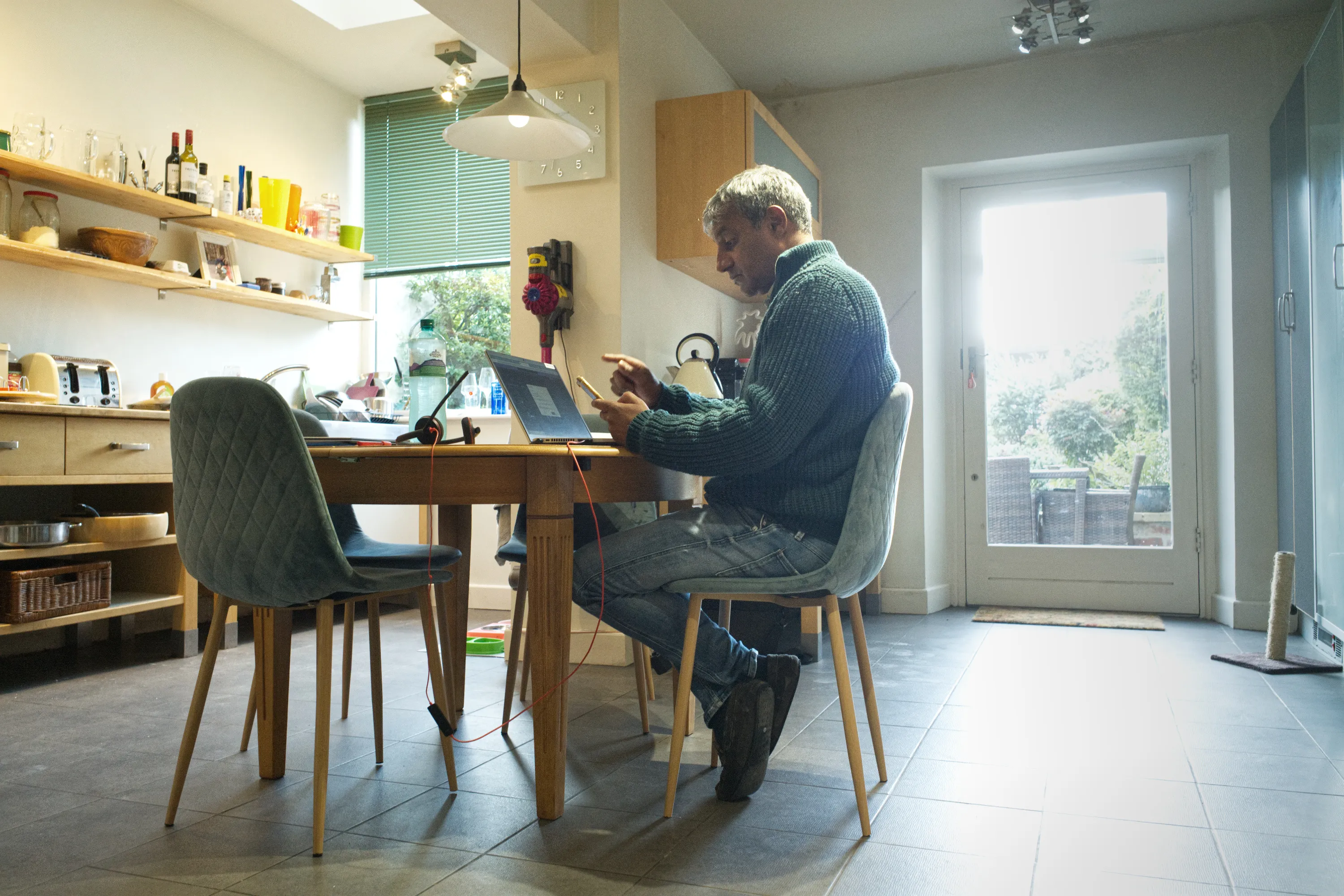 Man seated at a kitchen table with a laptop and tablet, garden door behind him