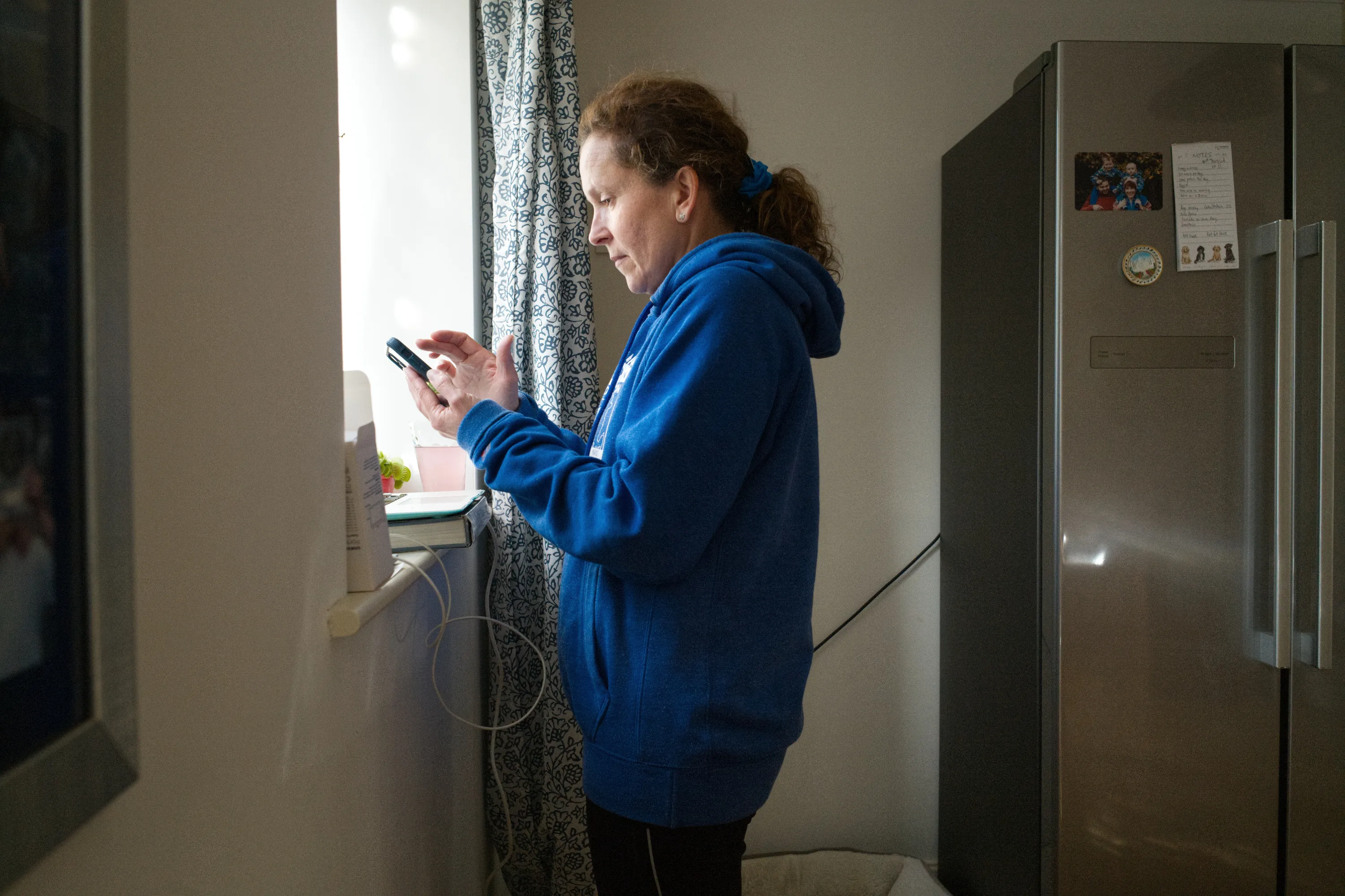 Woman in a blue hoodie looking at a phone by a kitchen window, fridge beside her