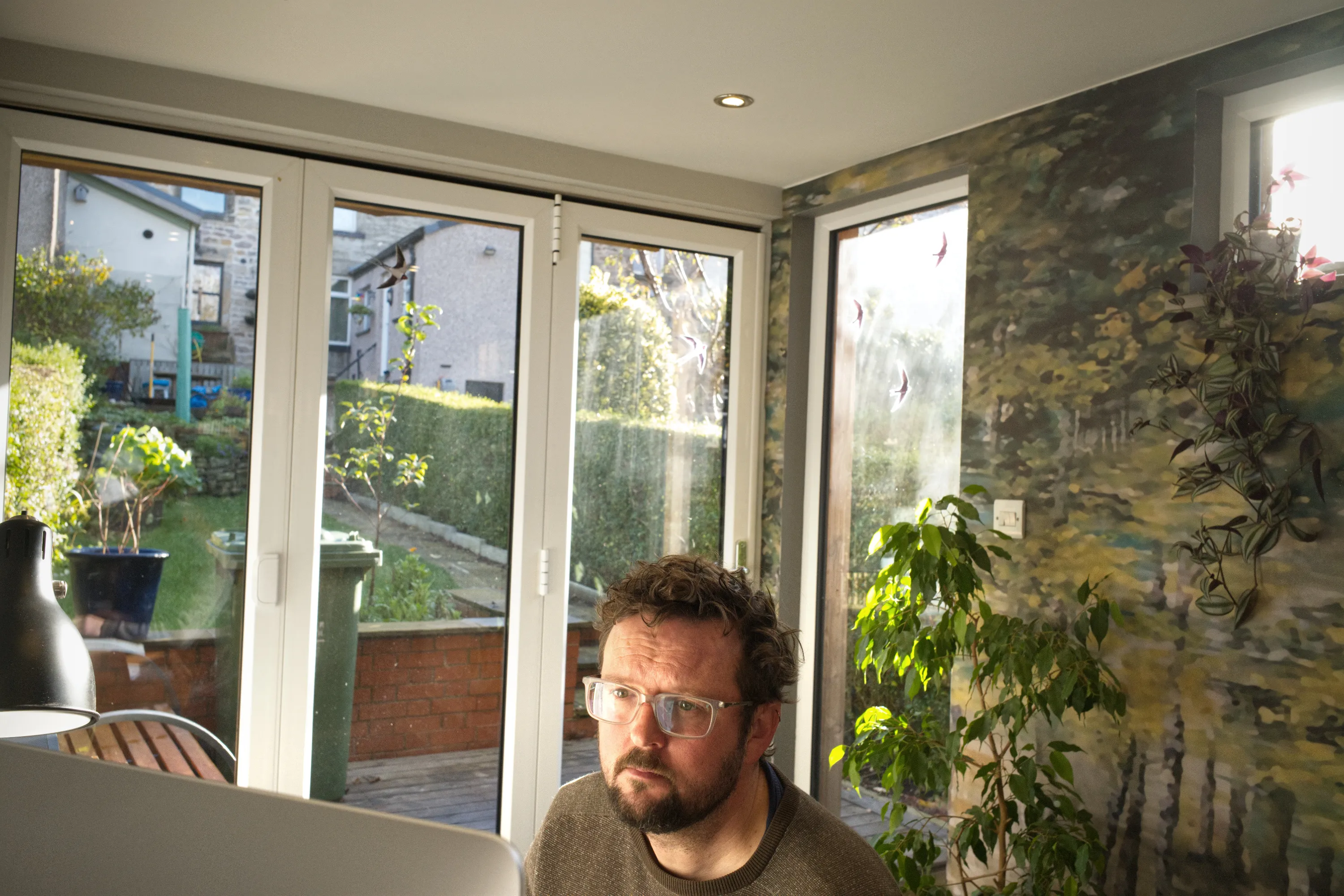 Bearded man at a laptop in a glass-walled extension, large mural on the wall behind him