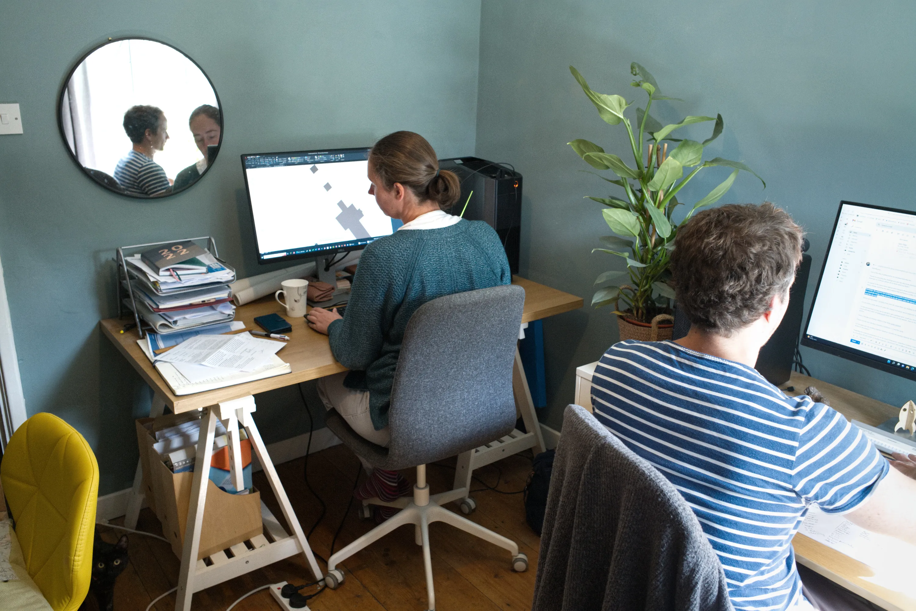 Two people working at side-by-side desks with monitors, round mirror on the wall