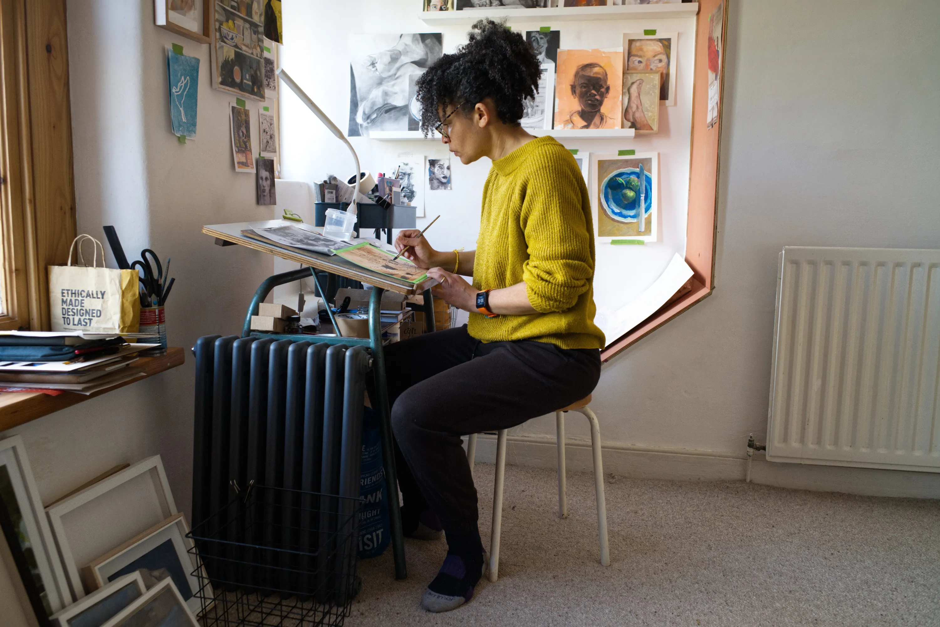 Woman painting at a drawing board in a studio surrounded by artworks on the wall