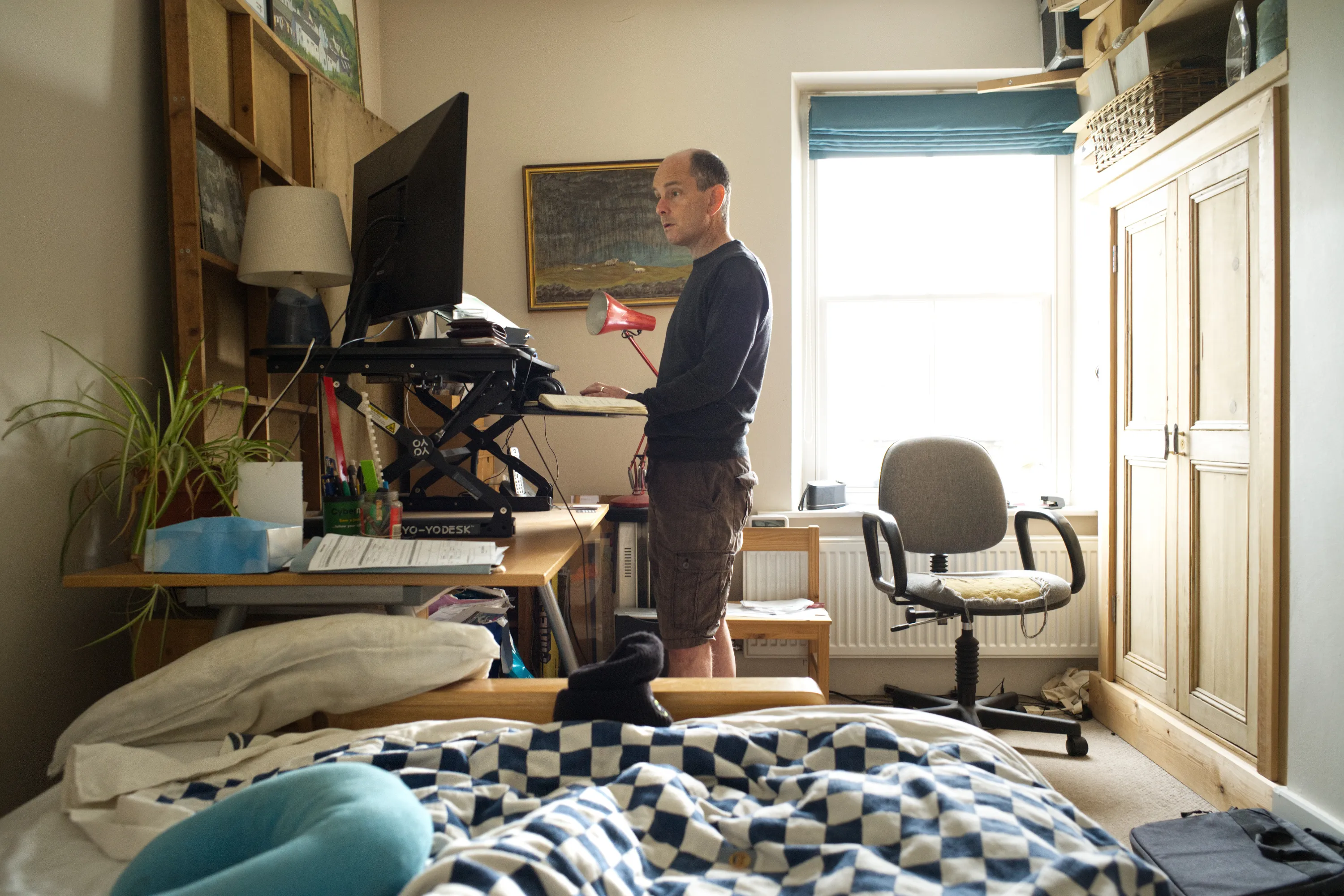 Man standing at a sit-stand desk with a large monitor in a bedroom