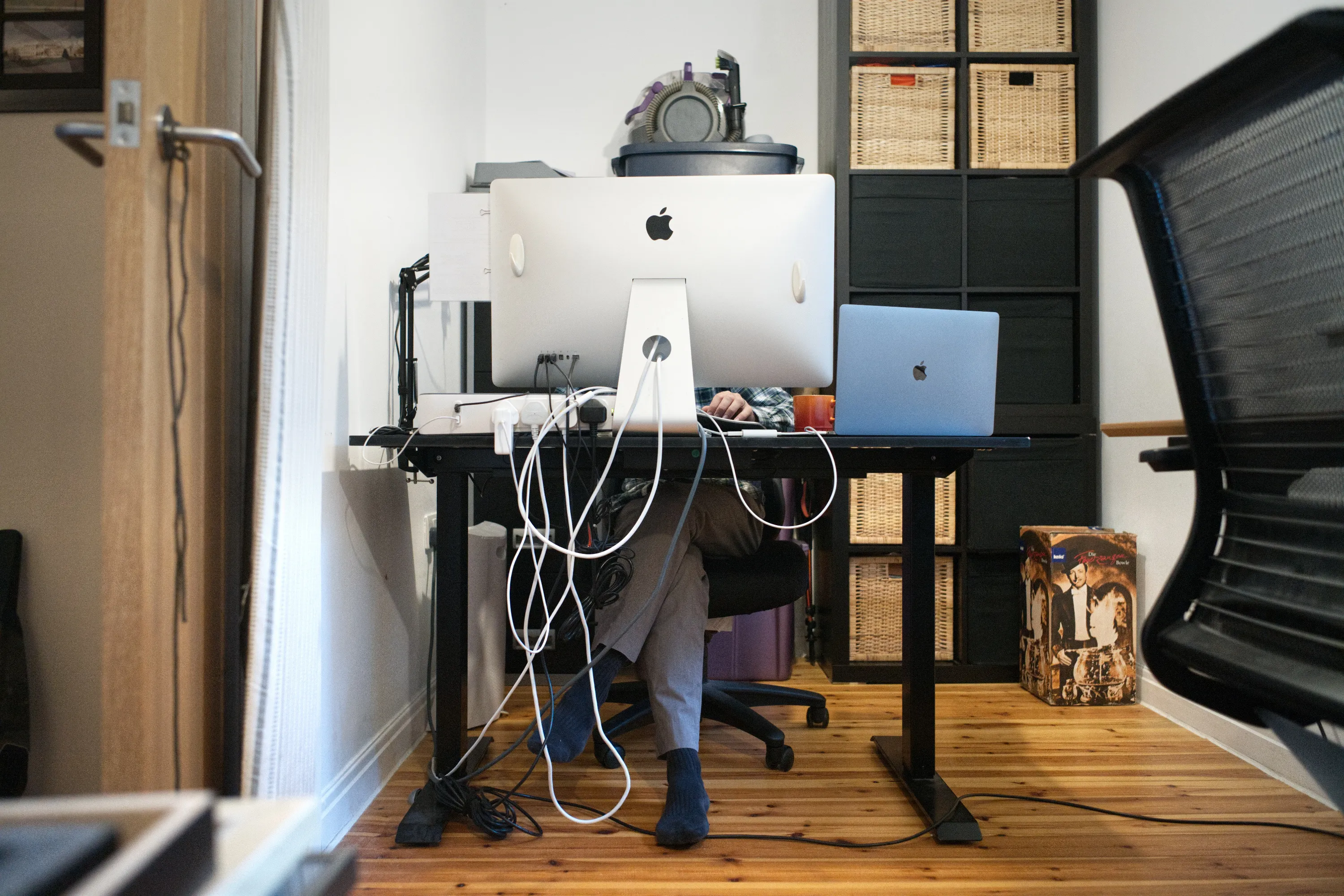 Person obscured behind an iMac and laptop on a height-adjustable desk, cables trailing to the floor