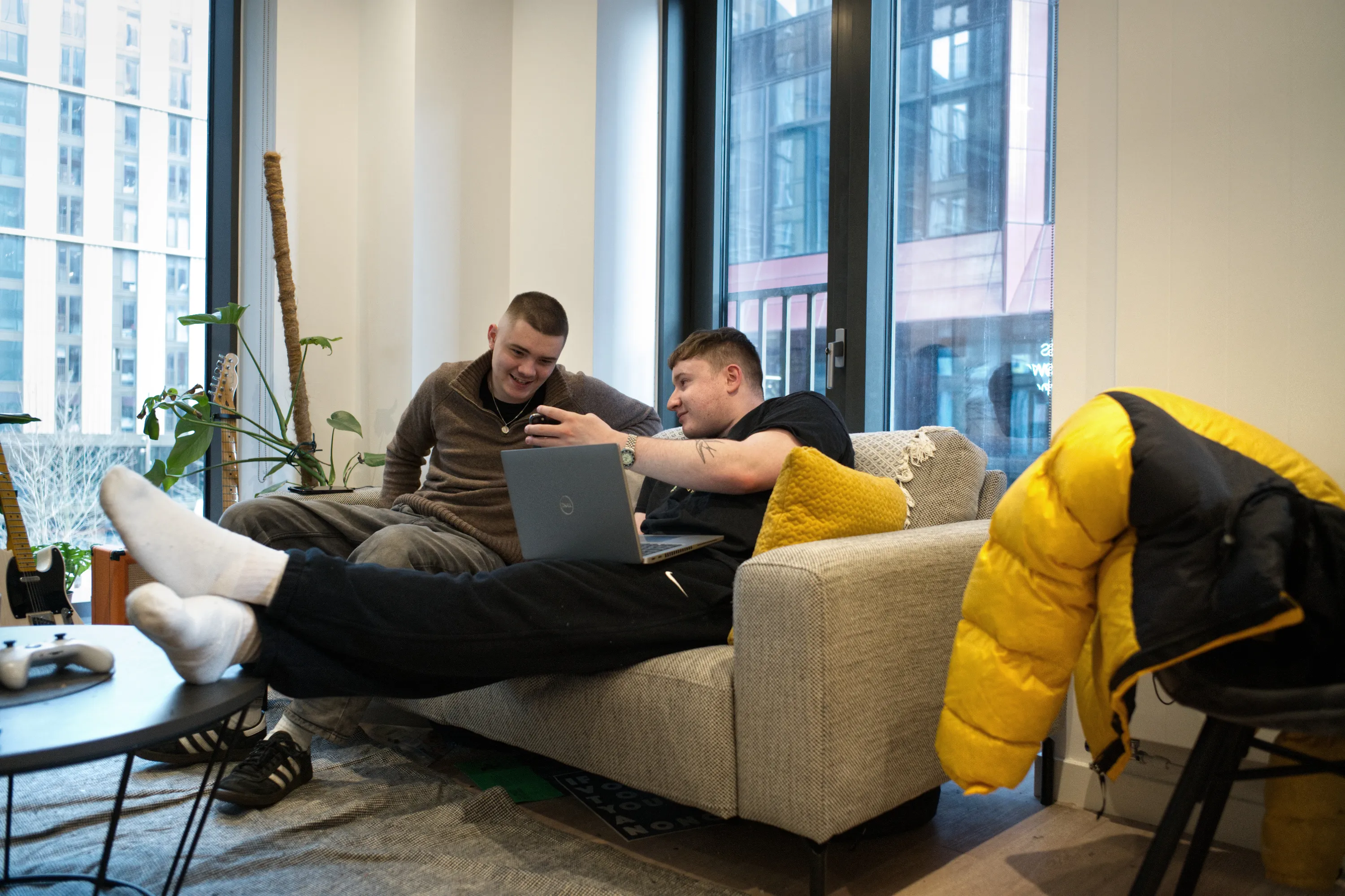 Two men on a sofa sharing a laptop in a bright apartment with floor-to-ceiling windows